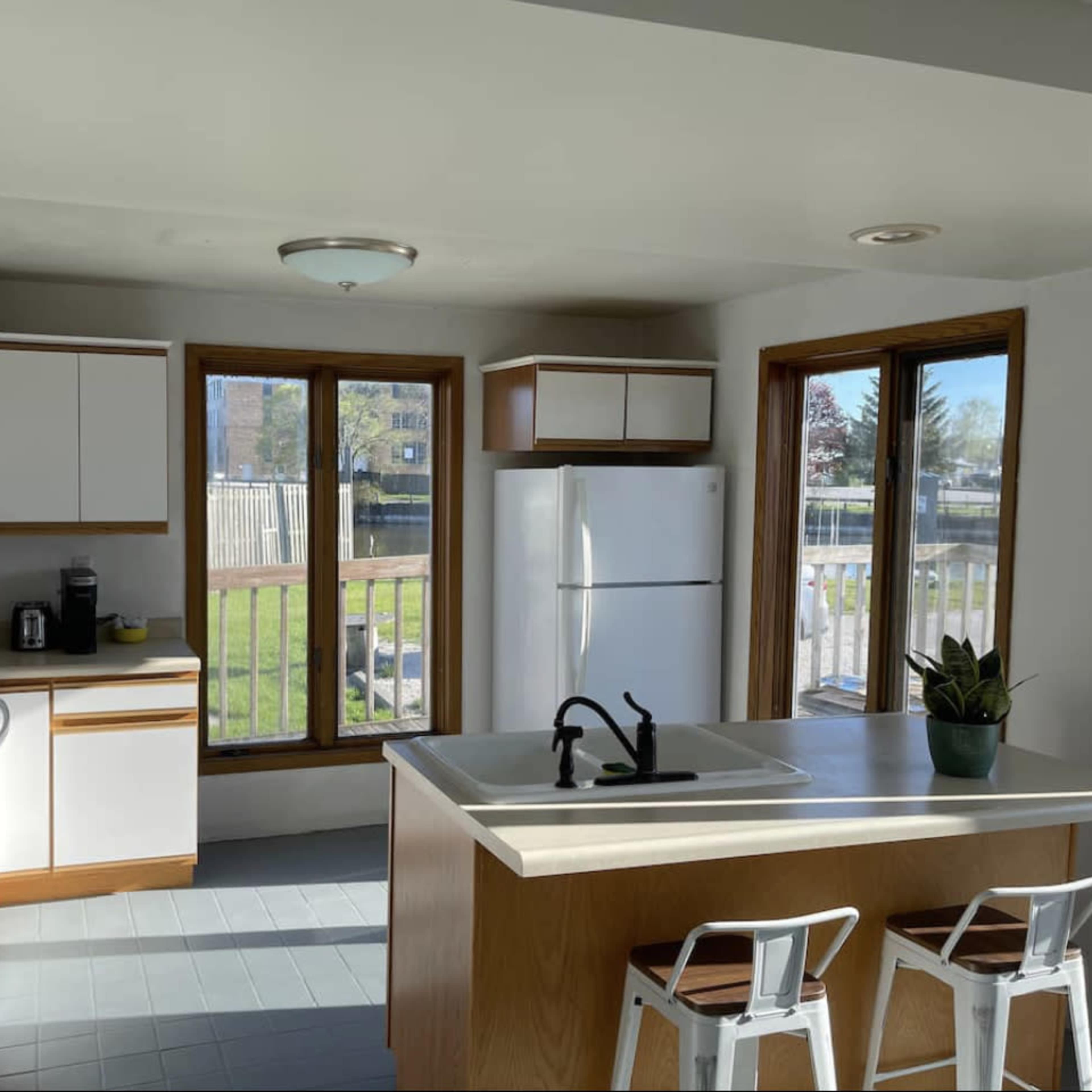 A bright kitchen with light-colored cabinetry, a white refrigerator, a sink on the island, and a view of the backyard through large windows.
