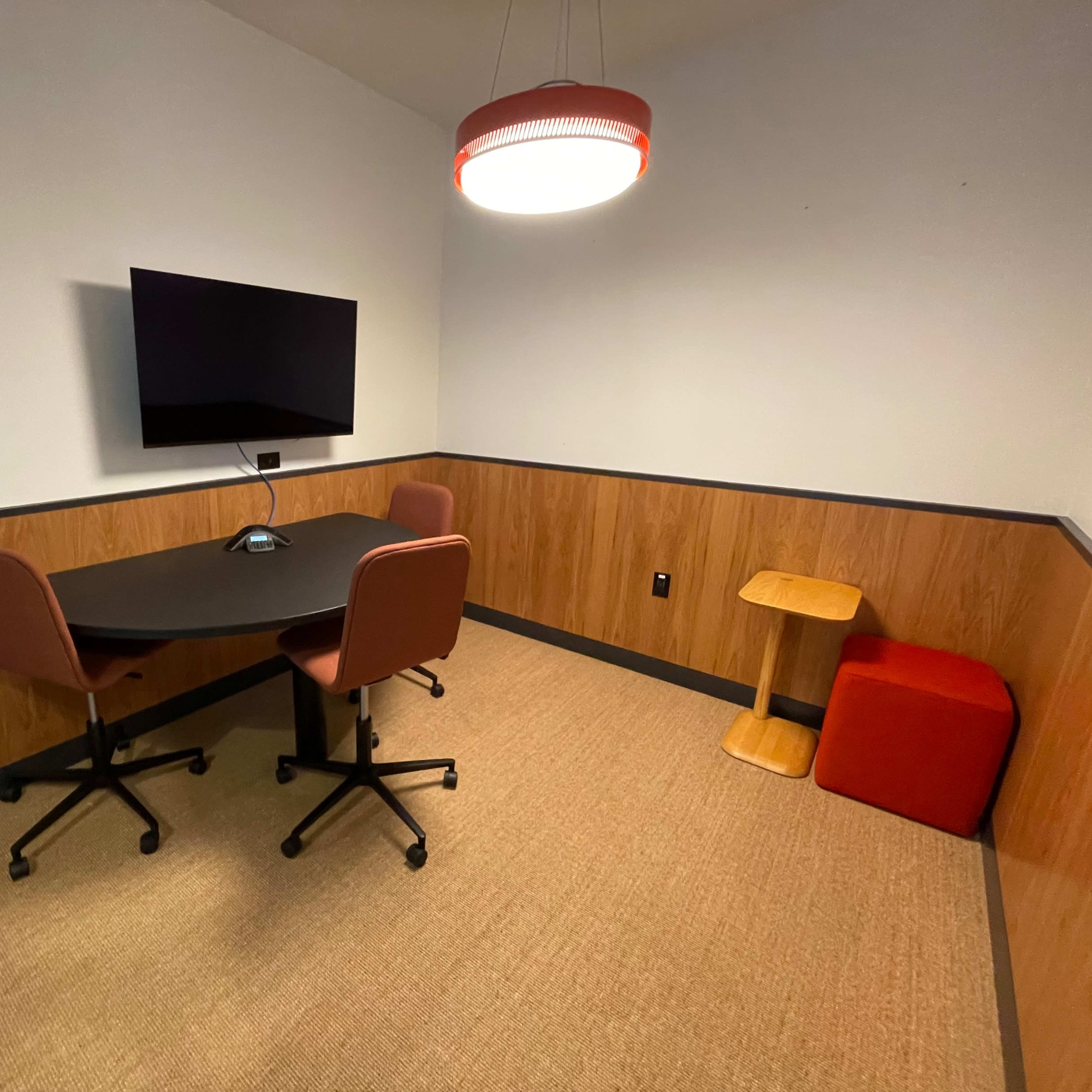 A small meeting room with a table, two chairs, a wall-mounted TV, and a side table, featuring wooden paneling and carpeted flooring.
