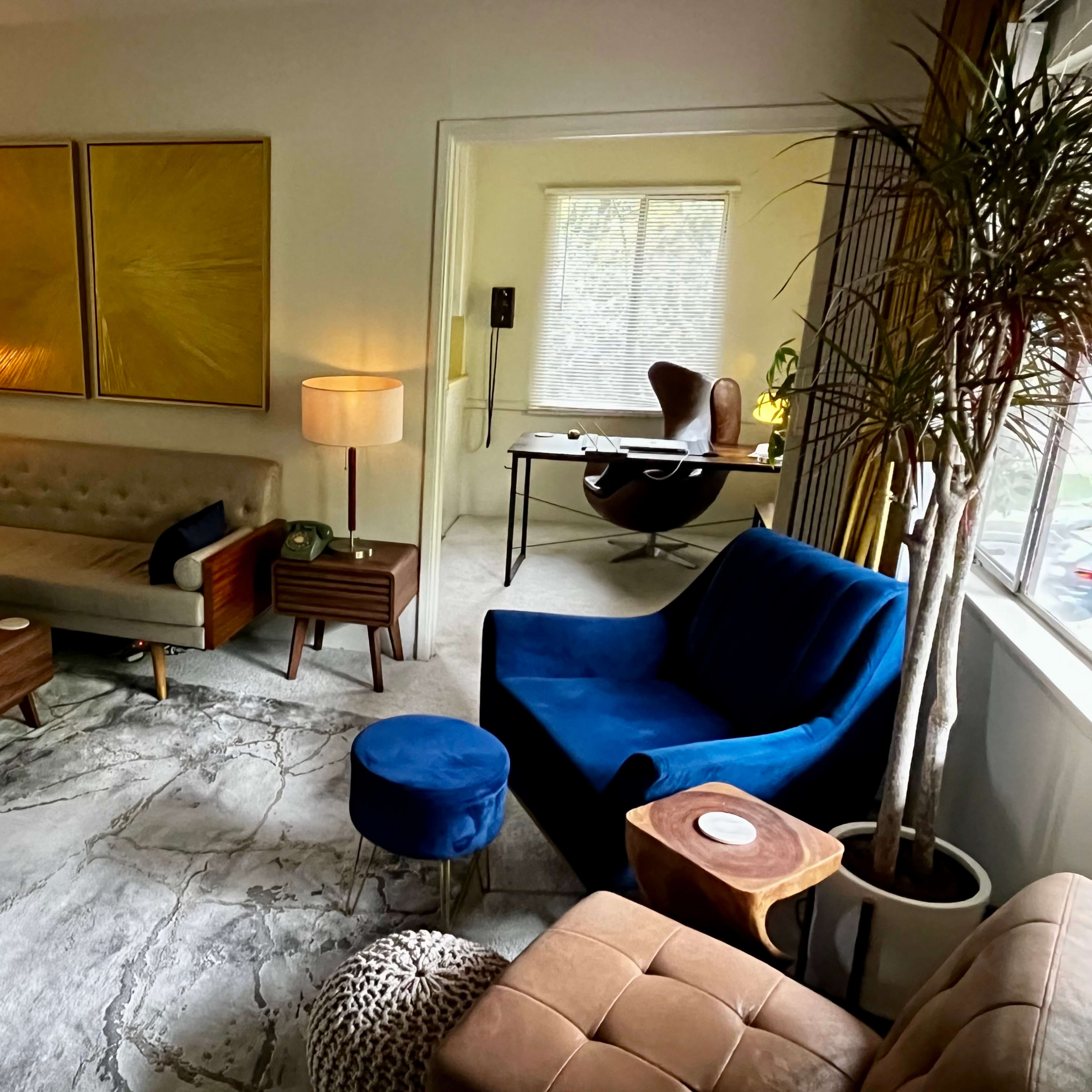 A stylish living space featuring a beige sofa, a blue velvet chair, a wooden coffee table, and a desk in an adjacent room.
