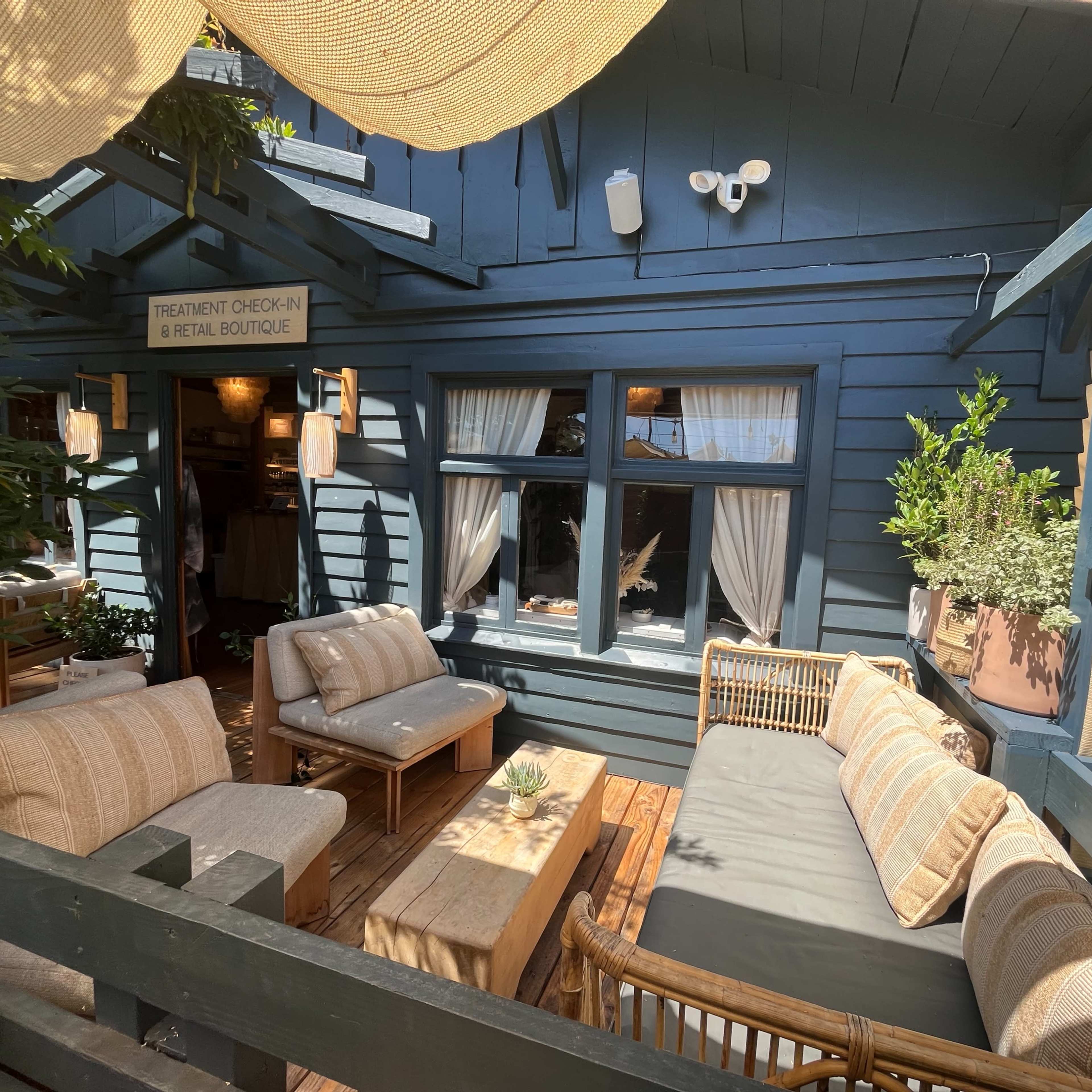 A cozy outdoor seating area with a wooden table and comfortable couches, adjacent to a blue building with large windows and greenery.