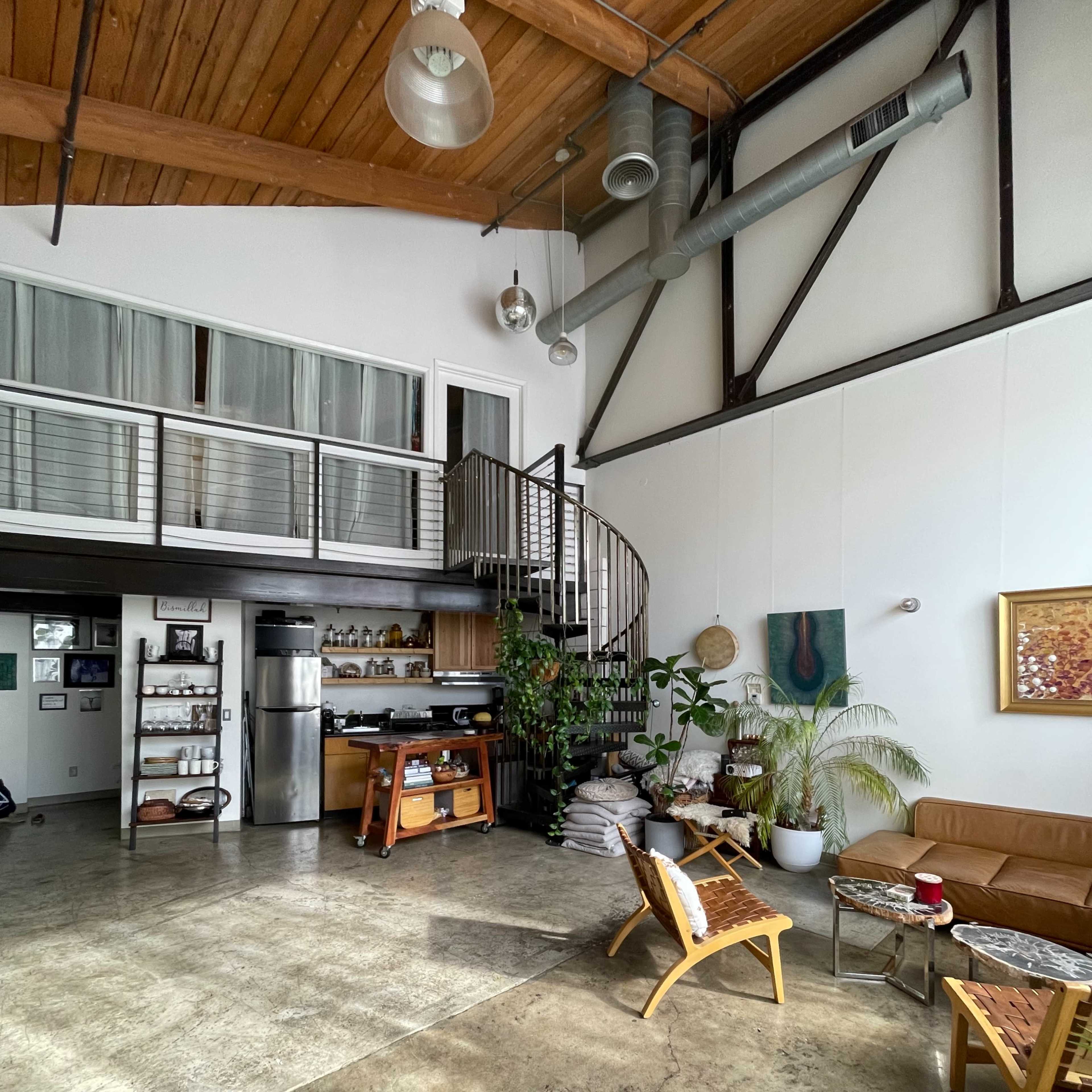 A spacious, industrial-style living area with high ceilings, a spiral staircase, and various plants arranged around natural stone flooring.