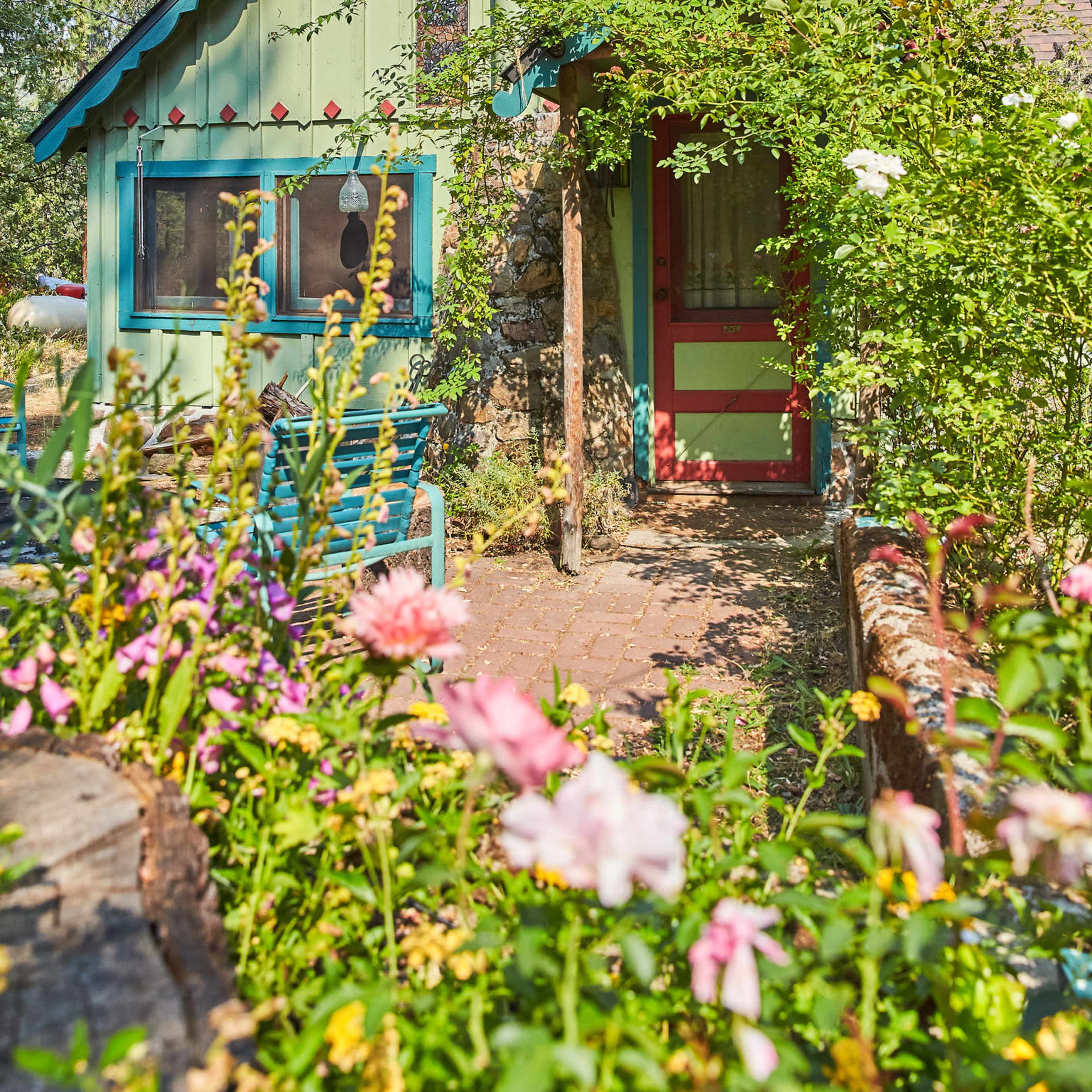 A small, brightly painted cottage is surrounded by colorful floral plants and greenery.