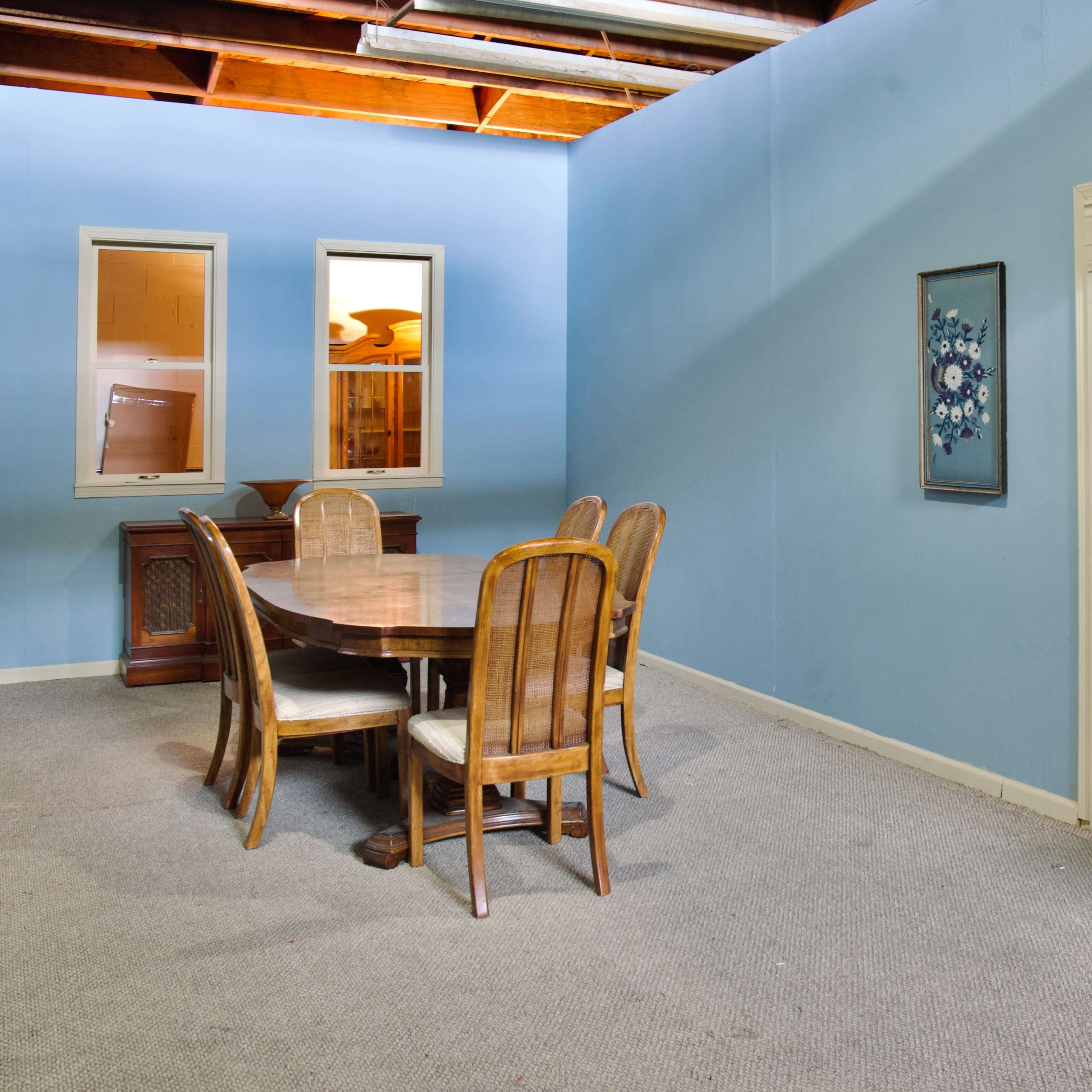 An empty room features a round wooden dining table surrounded by six chairs, with blue walls and a built-in shelf next to a small plant.