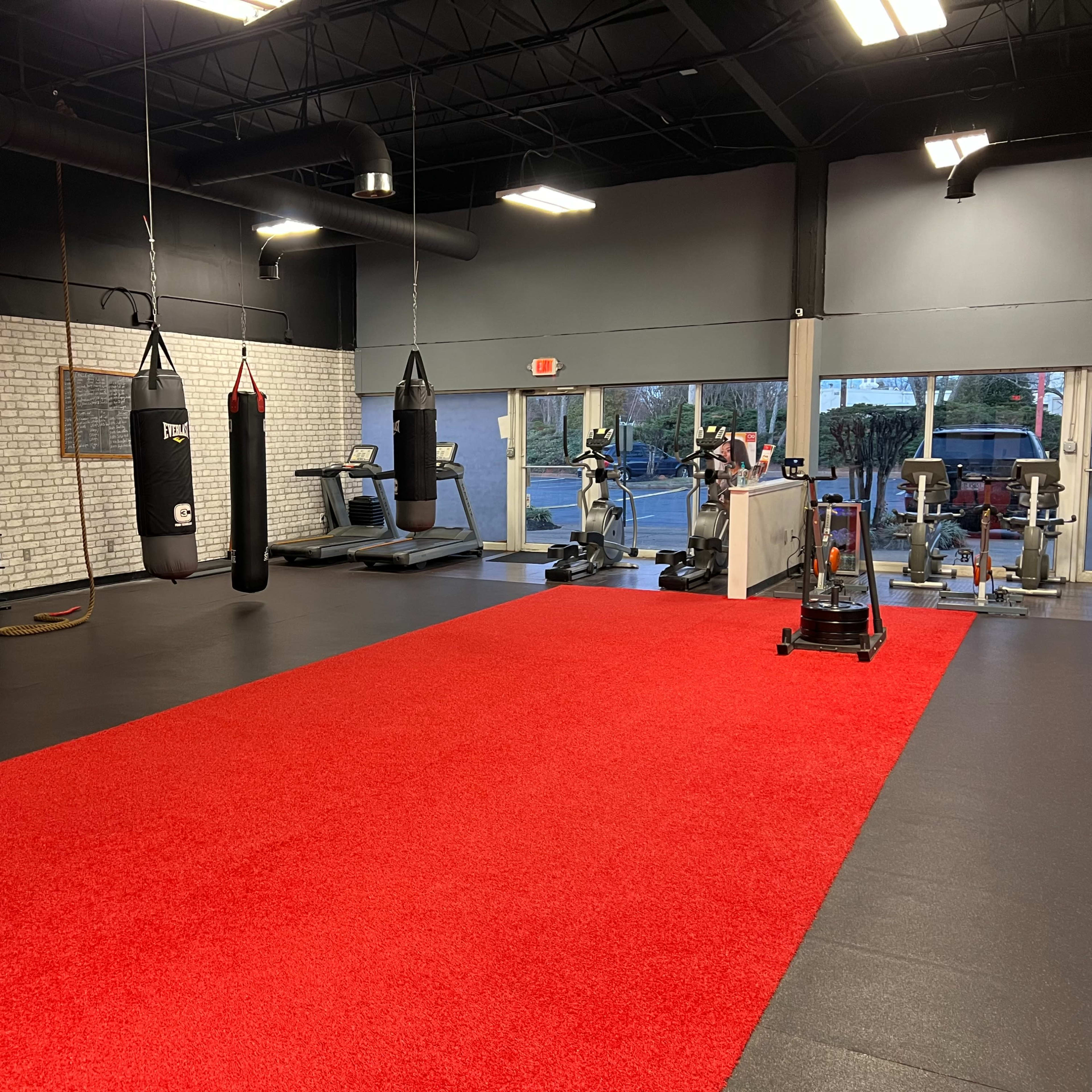 A spacious gym interior featuring red flooring, hanging punching bags, exercise equipment, and a reception area.