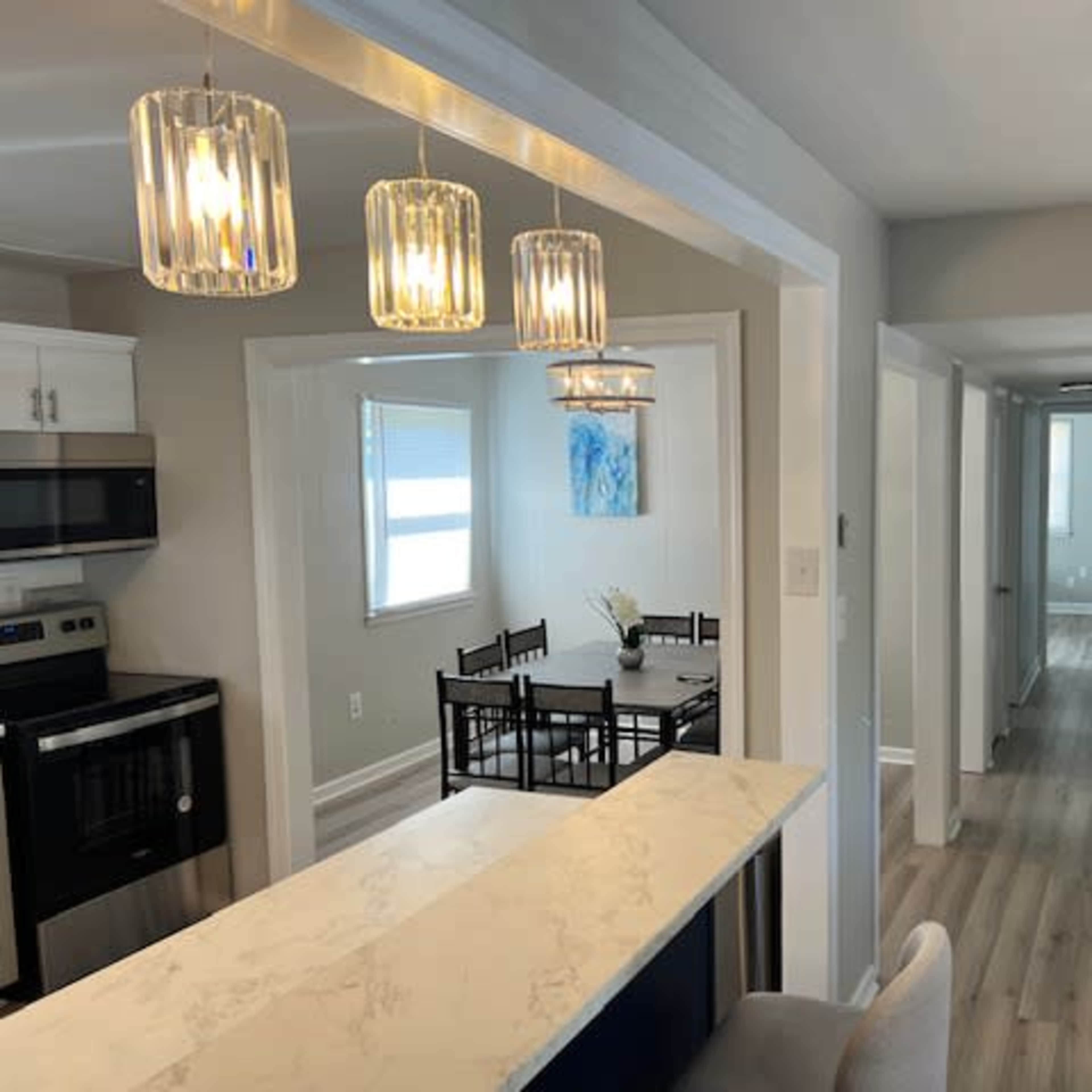 A modern, open-concept living space with a kitchen area featuring pendant lighting, a dining table in the adjacent room, and a hallway leading to more rooms.