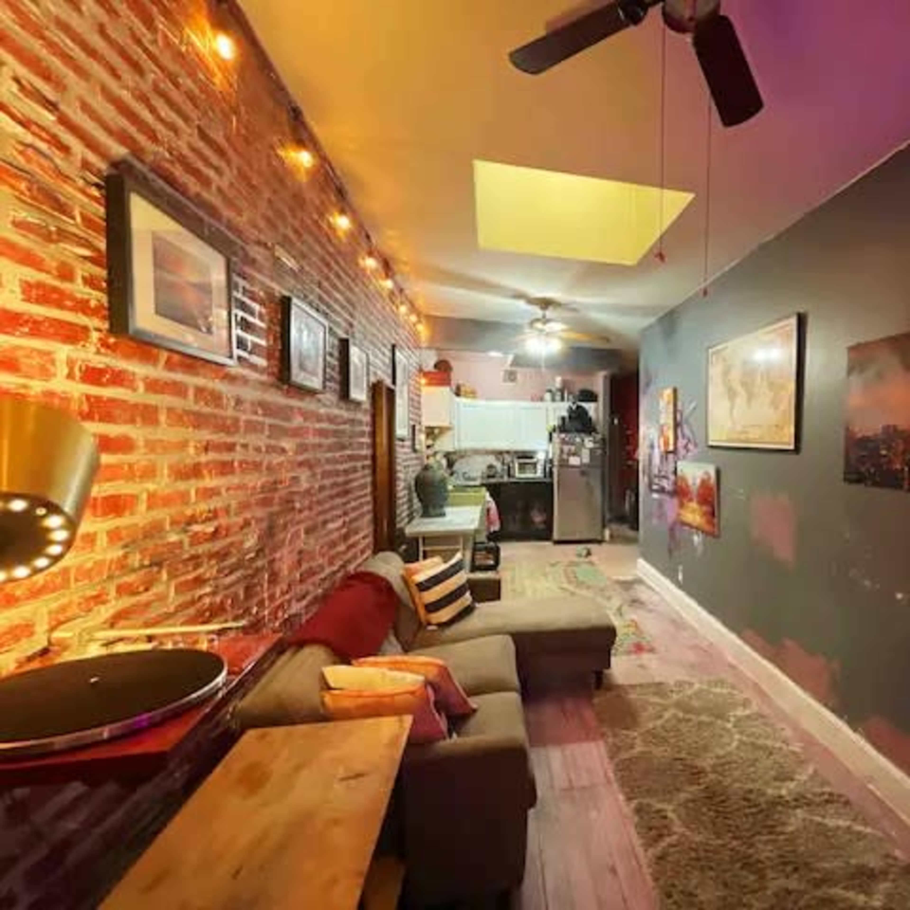 A narrow living area with exposed brick walls, a couch, and a small kitchen visible in the background.