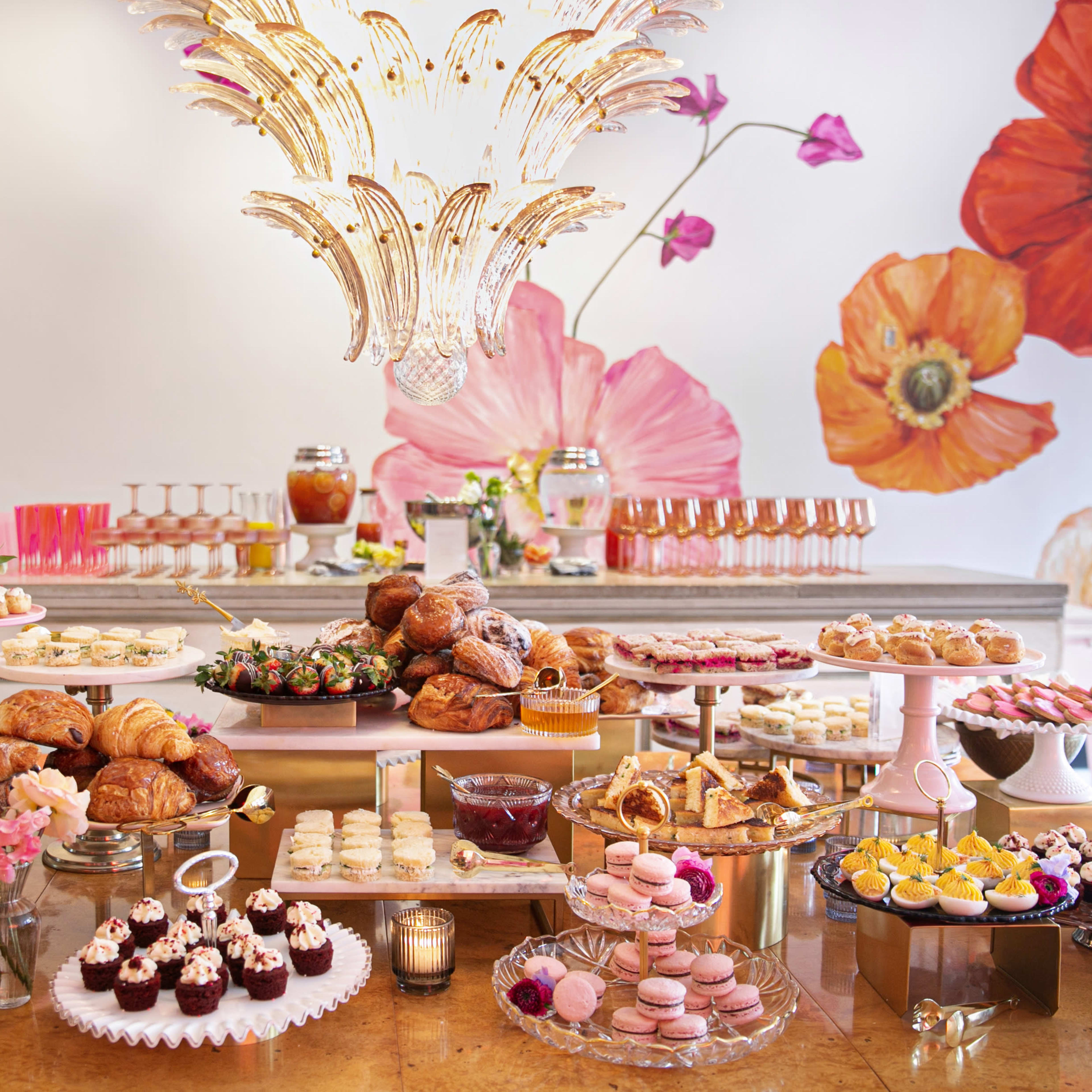 A lavish dessert table is arranged with an assortment of pastries, cakes, and decorative drinks beneath a floral mural.