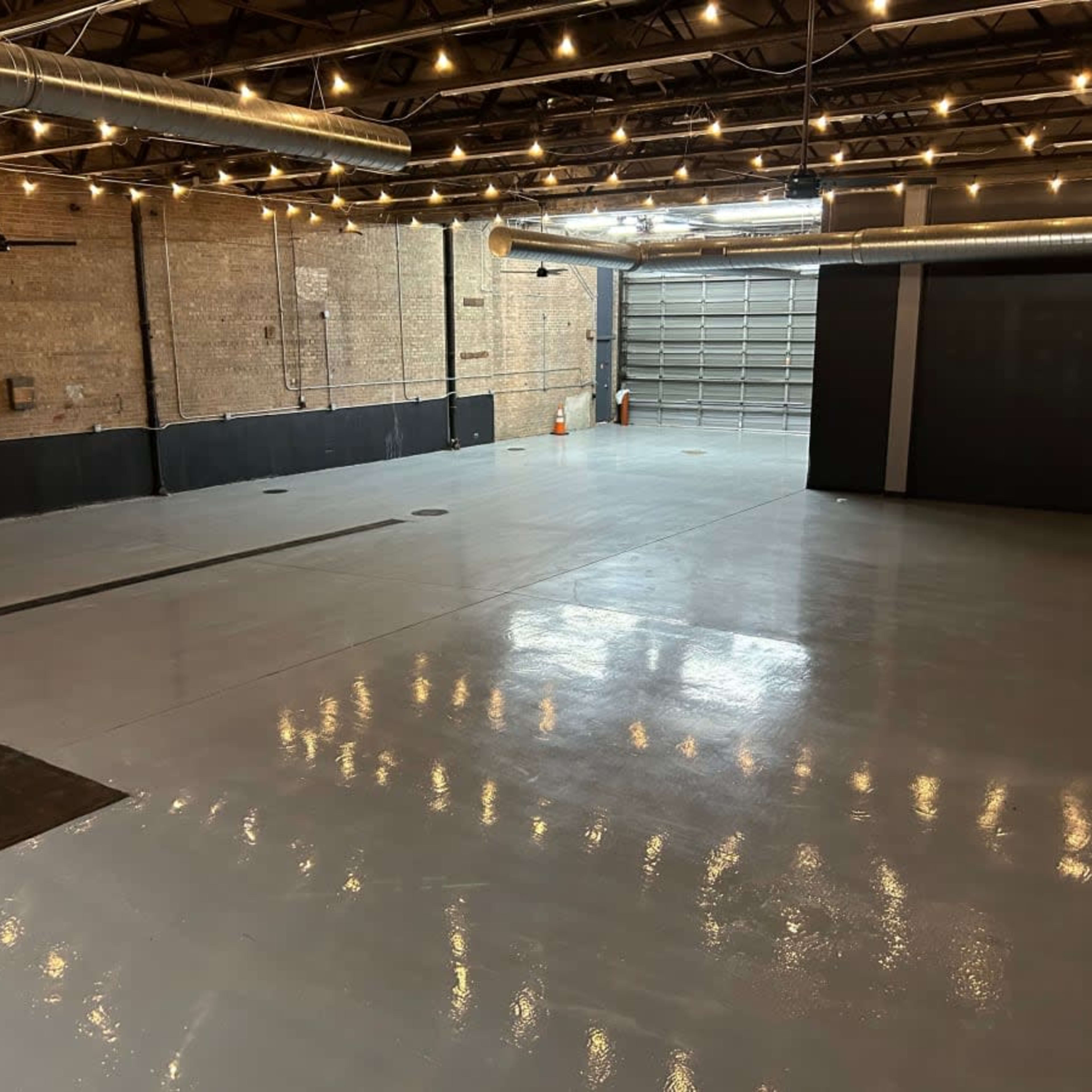 A large, empty industrial space with a polished gray concrete floor and exposed brick walls, featuring overhead lighting and a garage door.