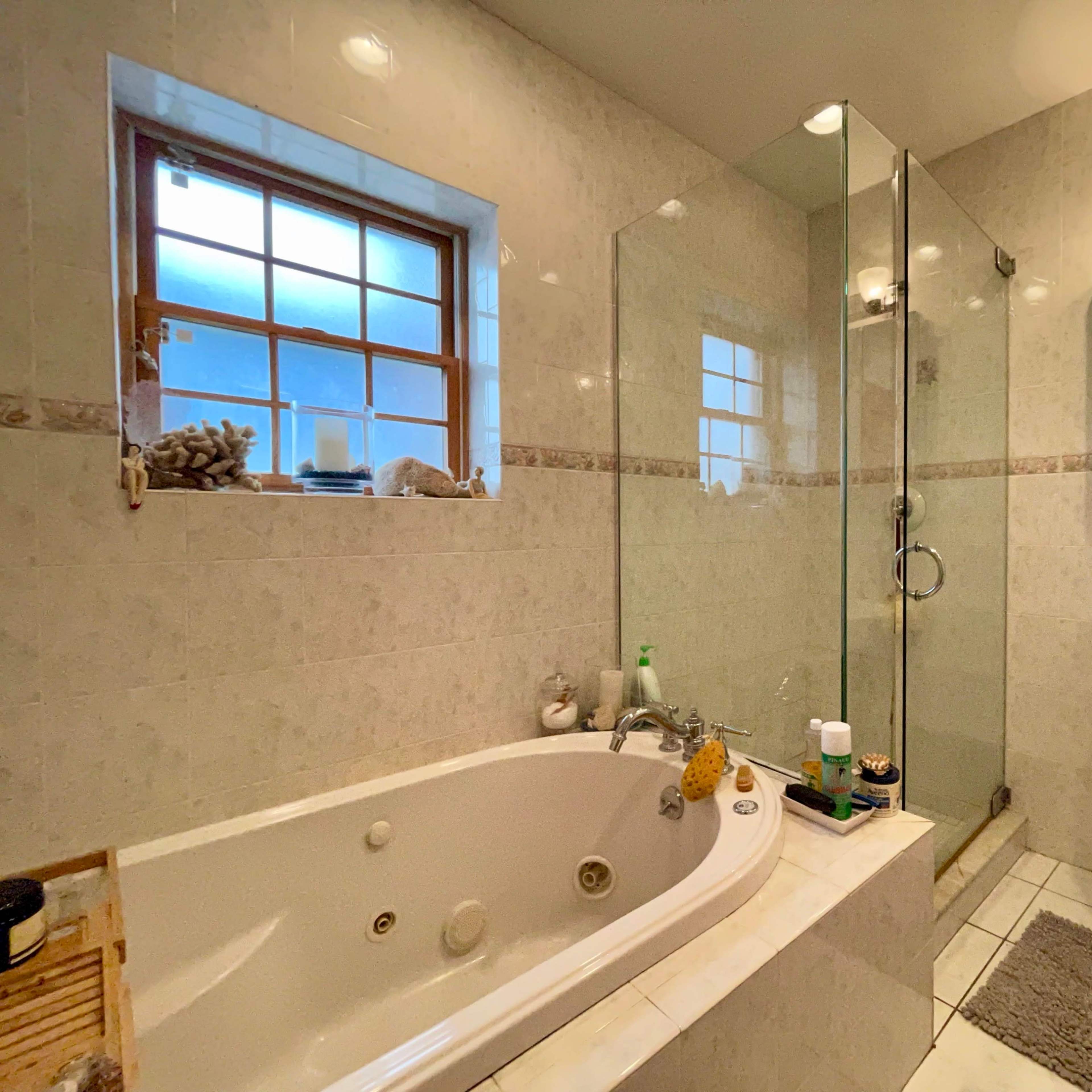 The bathroom features a glass shower enclosure, a corner soaking tub, a pedestal sink, and a window allowing natural light.