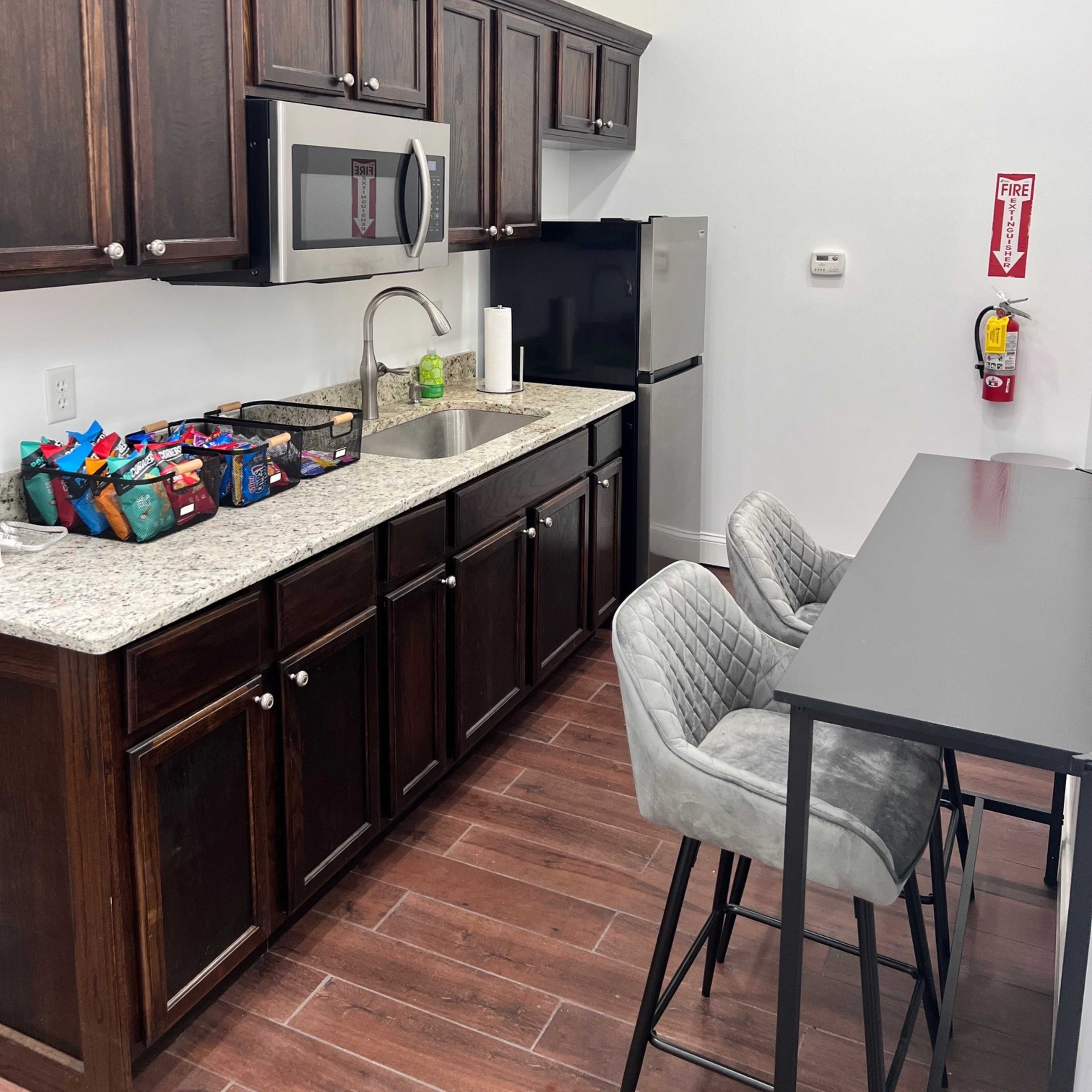 A modern kitchenette with dark wood cabinets, a stainless steel microwave, a small refrigerator, and a black table with gray chairs.