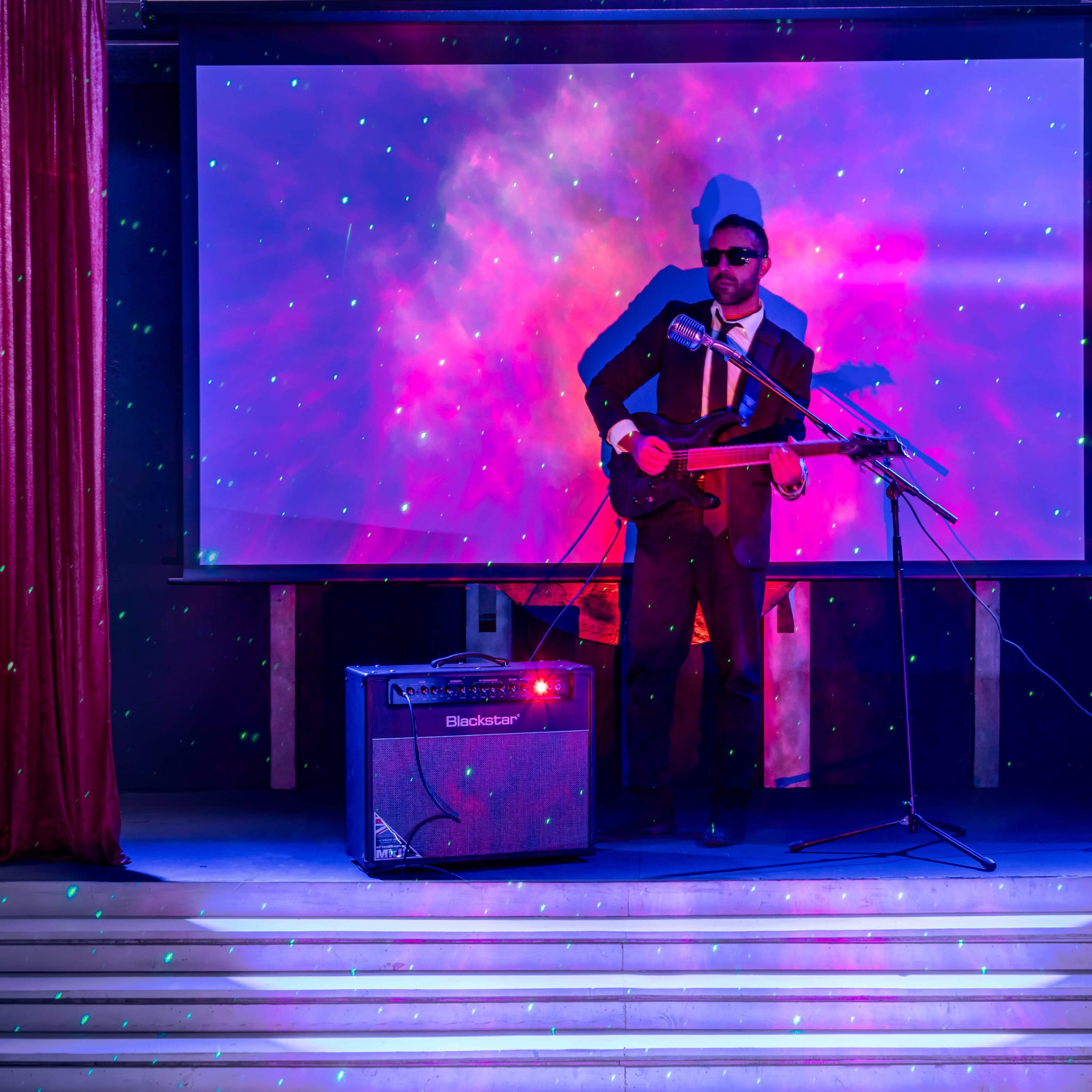 A musician in a suit plays guitar on stage in front of a colorful, illuminated backdrop.