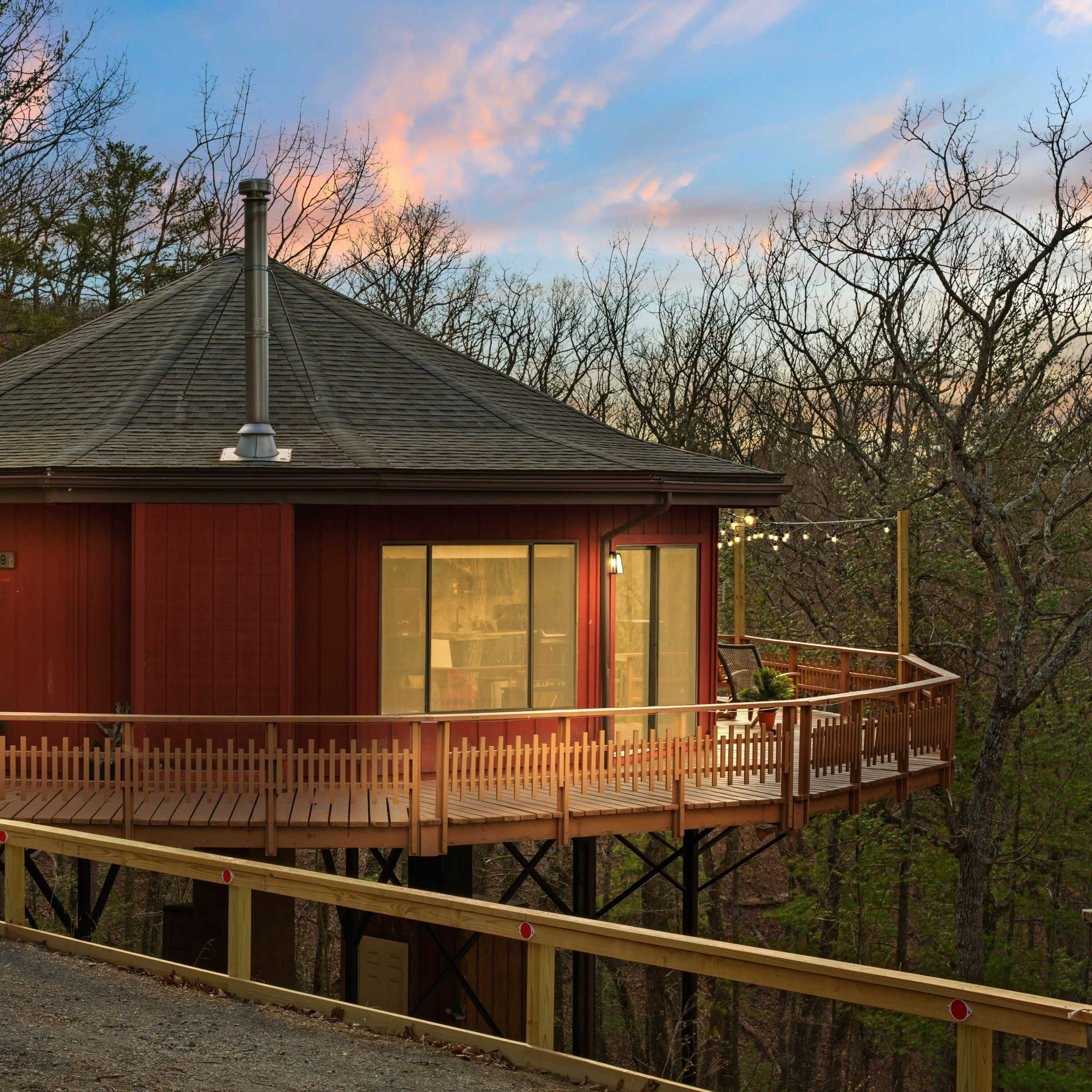 A round red cabin on stilts is surrounded by trees, with a deck that wraps around its exterior and a sunset sky in the background.