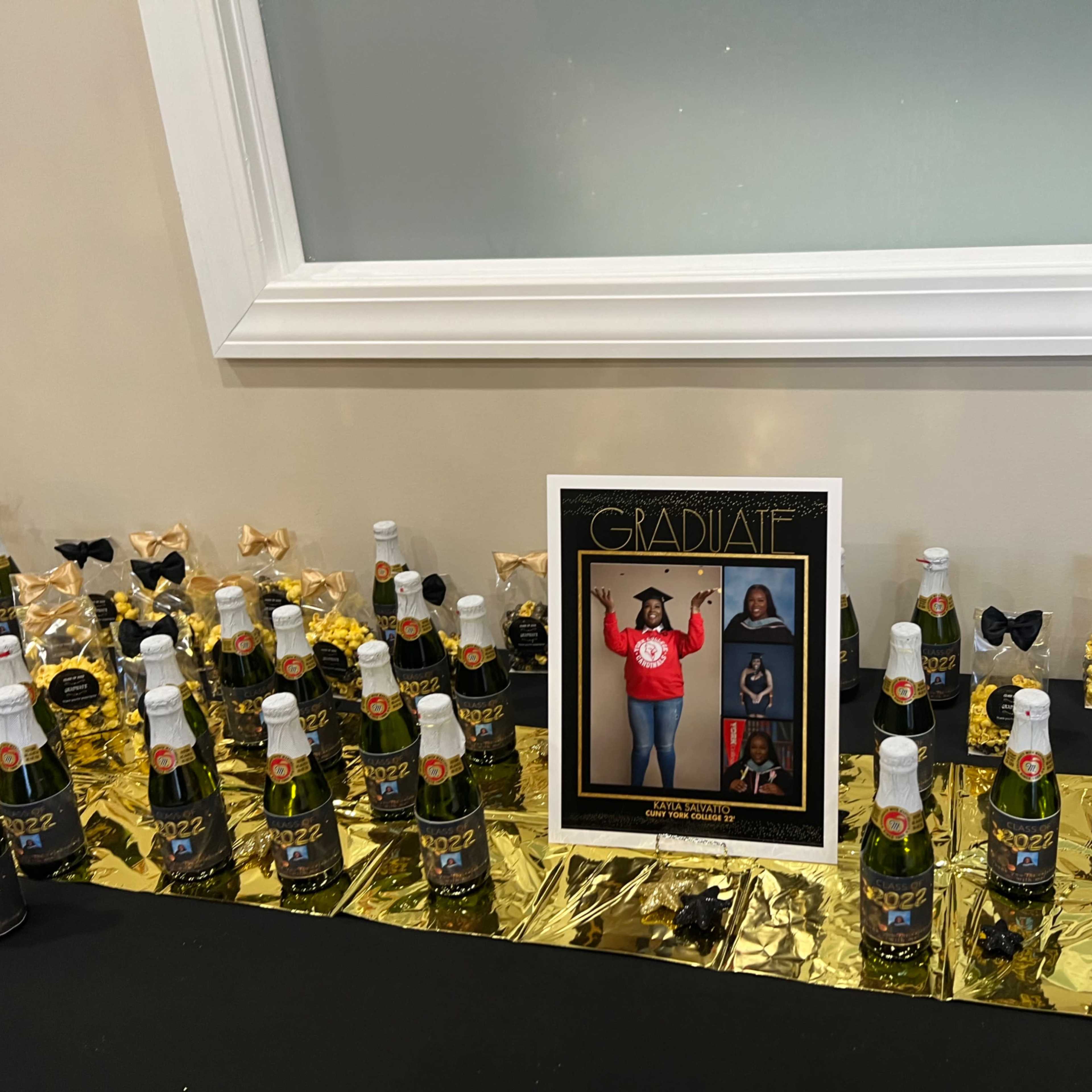 A table is adorned with decorated bottles of sparkling beverage, gold foil, and a framed graduation photo display.