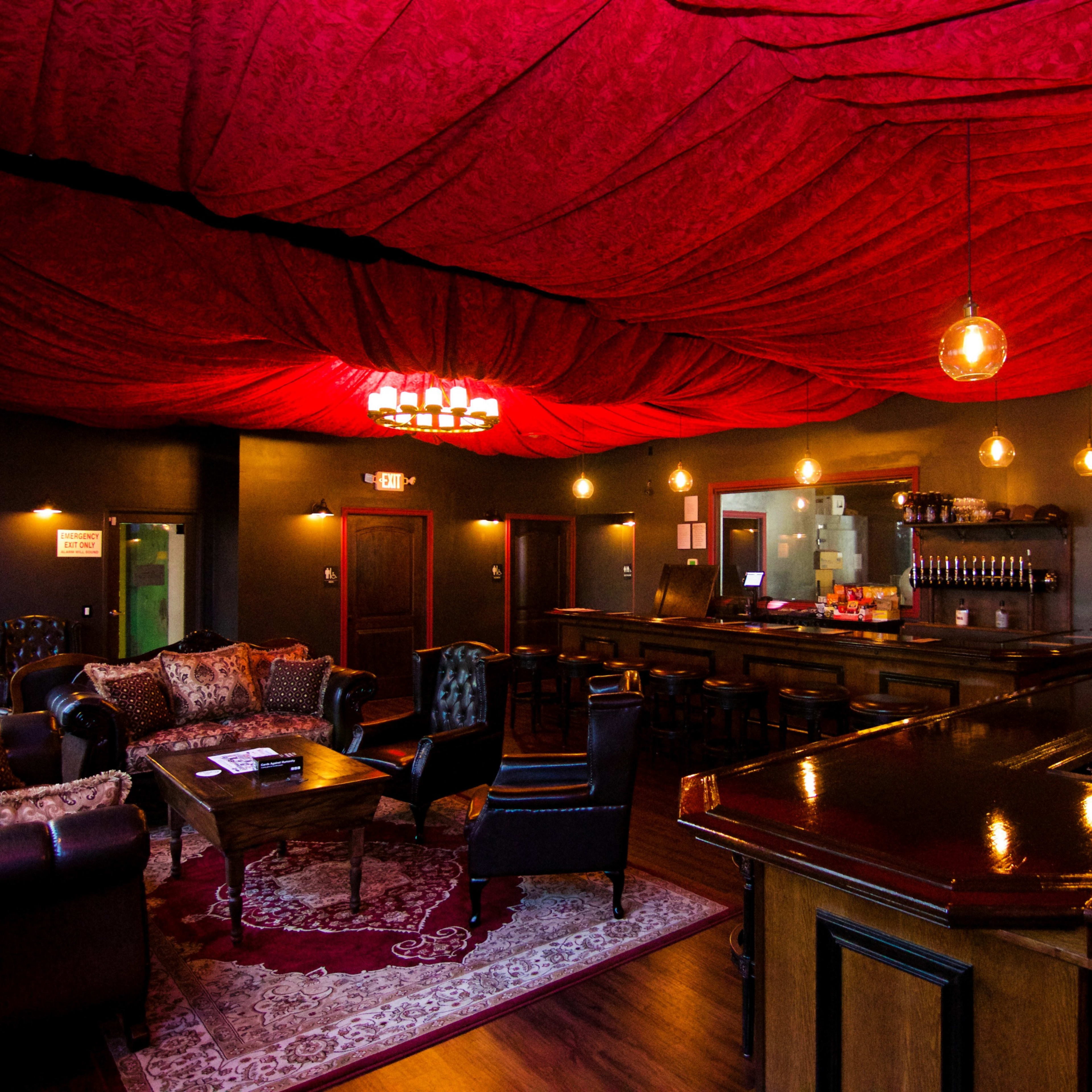 A cozy lounge with a red draped ceiling, a bar area, comfortable seating, and warm pendant lights.