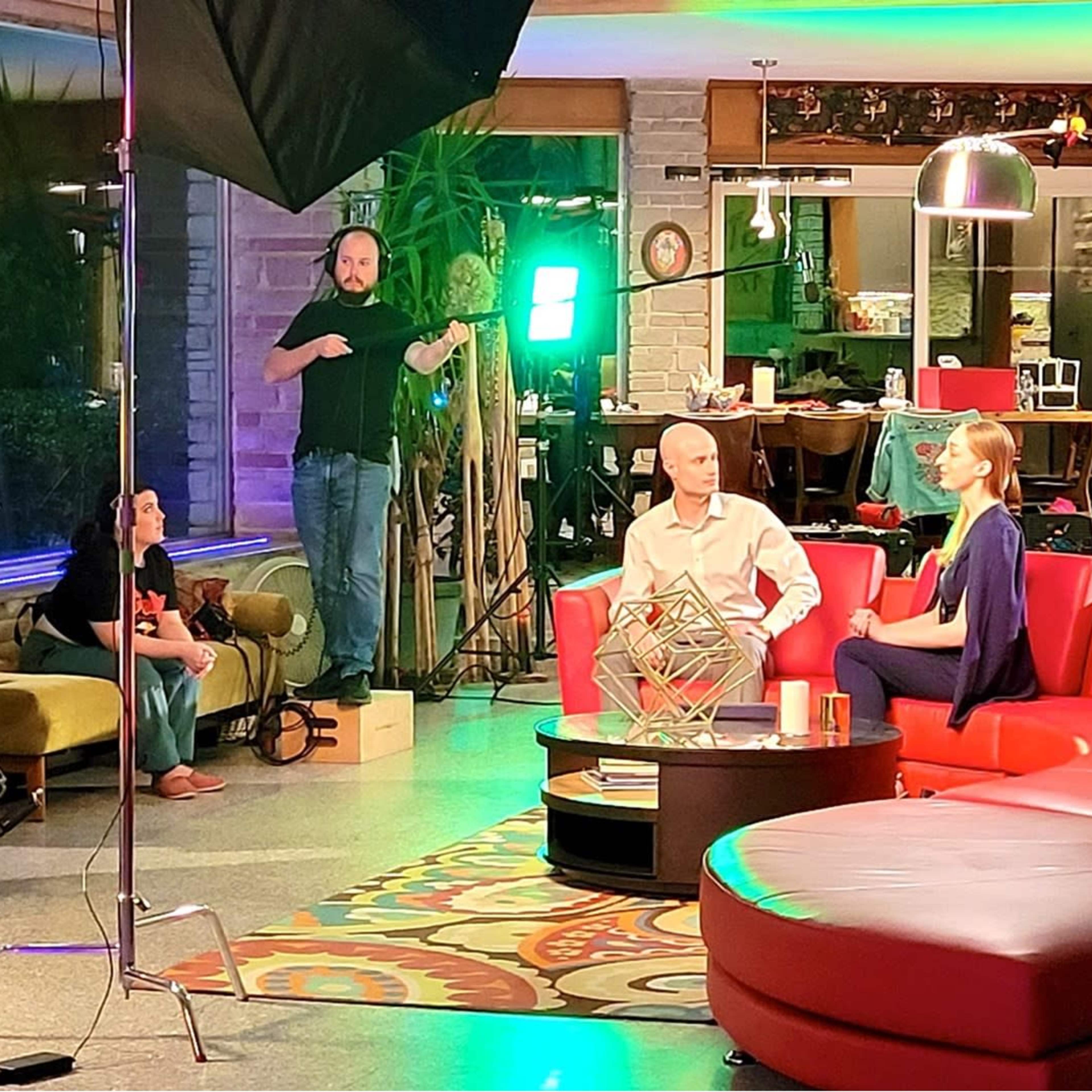 A film crew is setting up equipment for an interview scene in a modern indoor space featuring colorful furniture and decorative lighting.