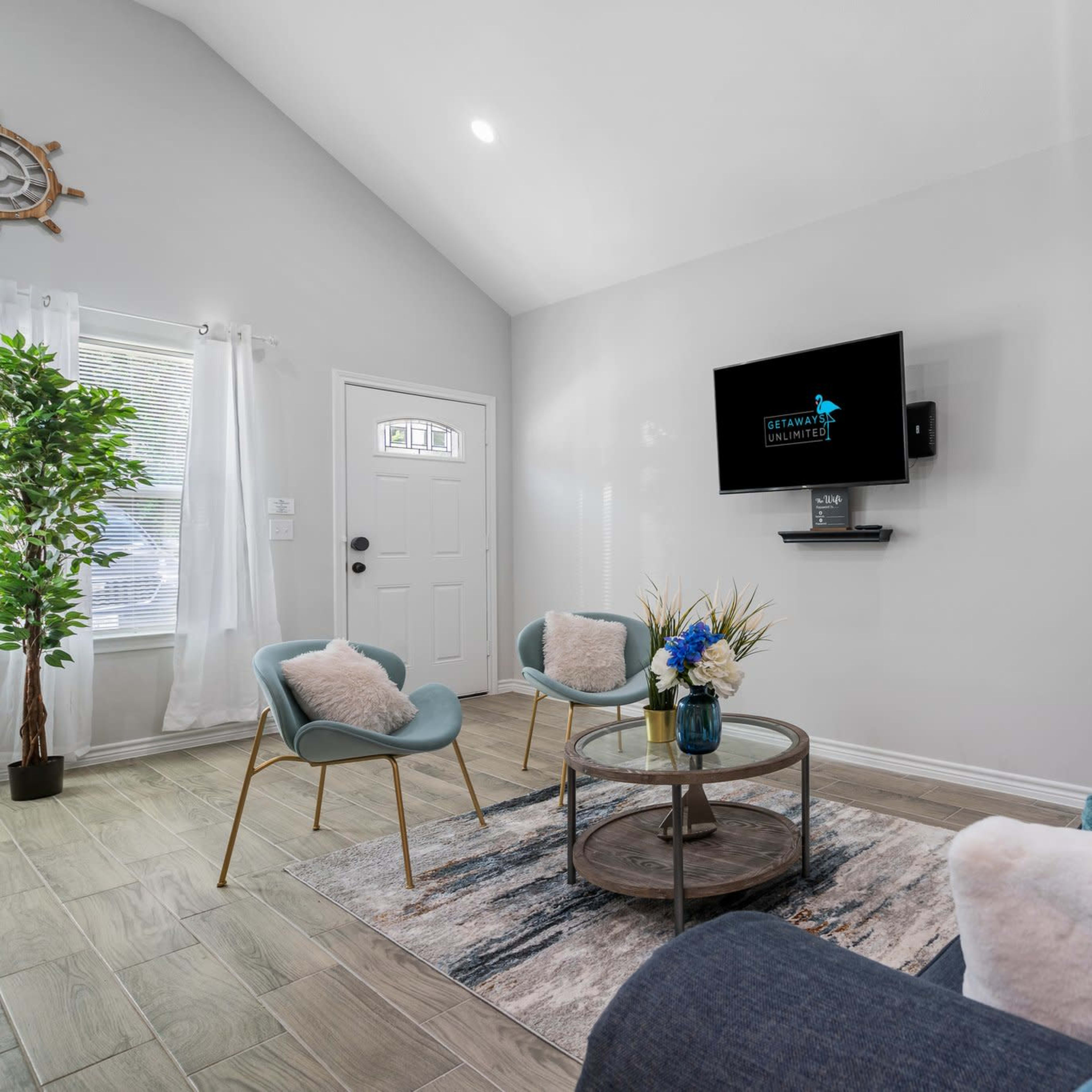 The living room features a blue sofa, two light blue chairs, a round coffee table with a vase of flowers, and a mounted television on a light gray wall.