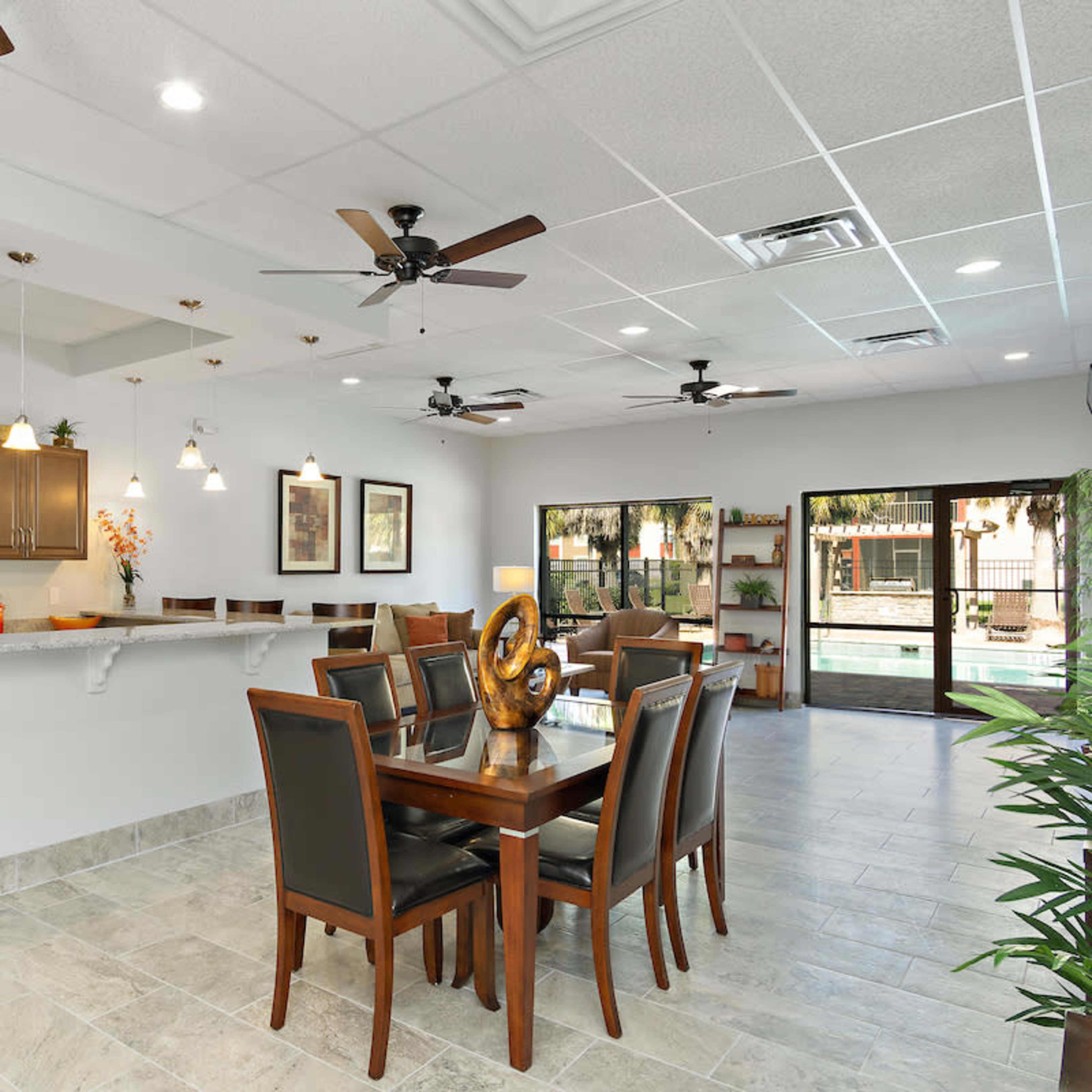 A spacious dining area features a large wooden table with chairs, a kitchen with dark cabinetry, and large windows that overlook a pool area.