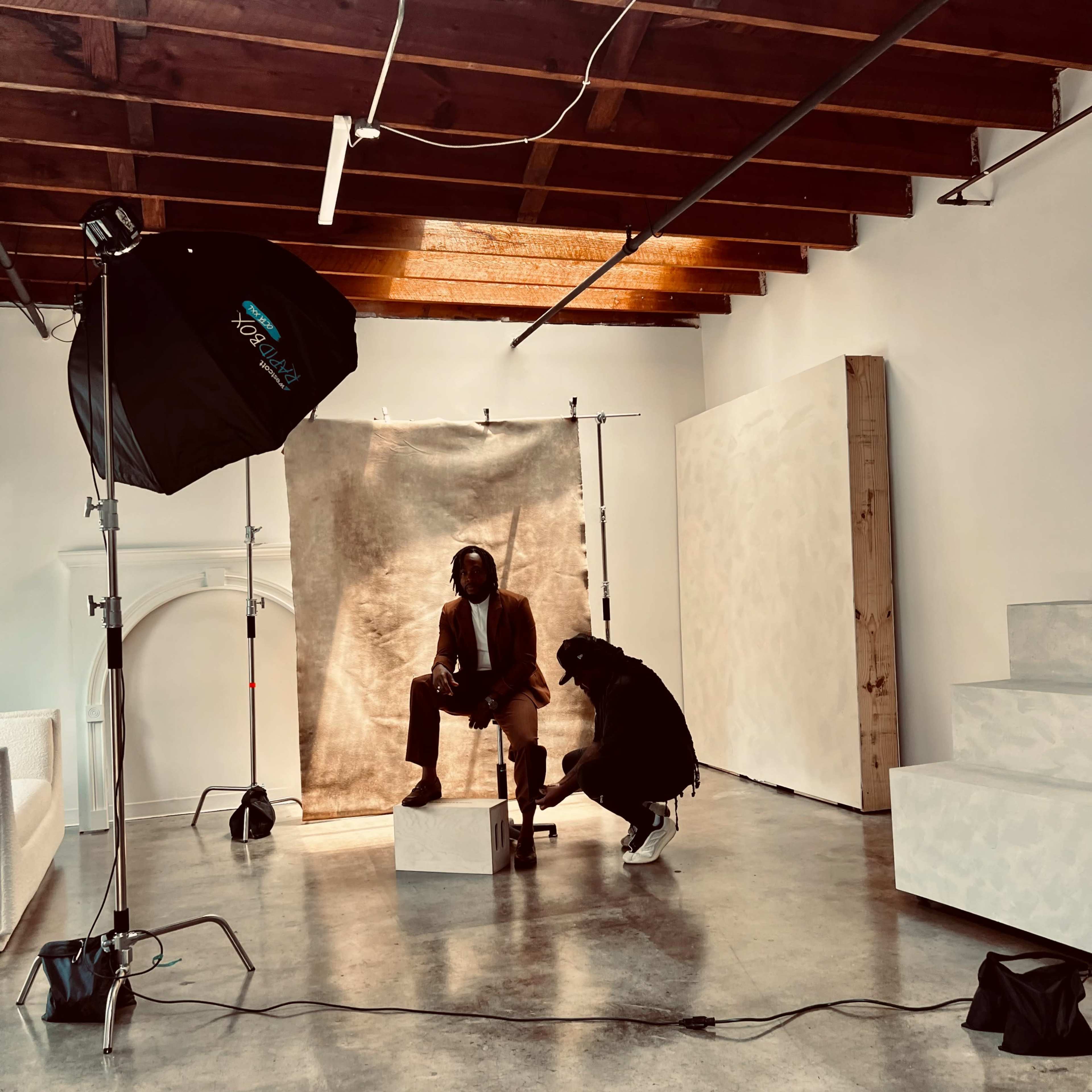 A model sits on a pedestal in a photo studio while a photographer adjusts the model's footwear.