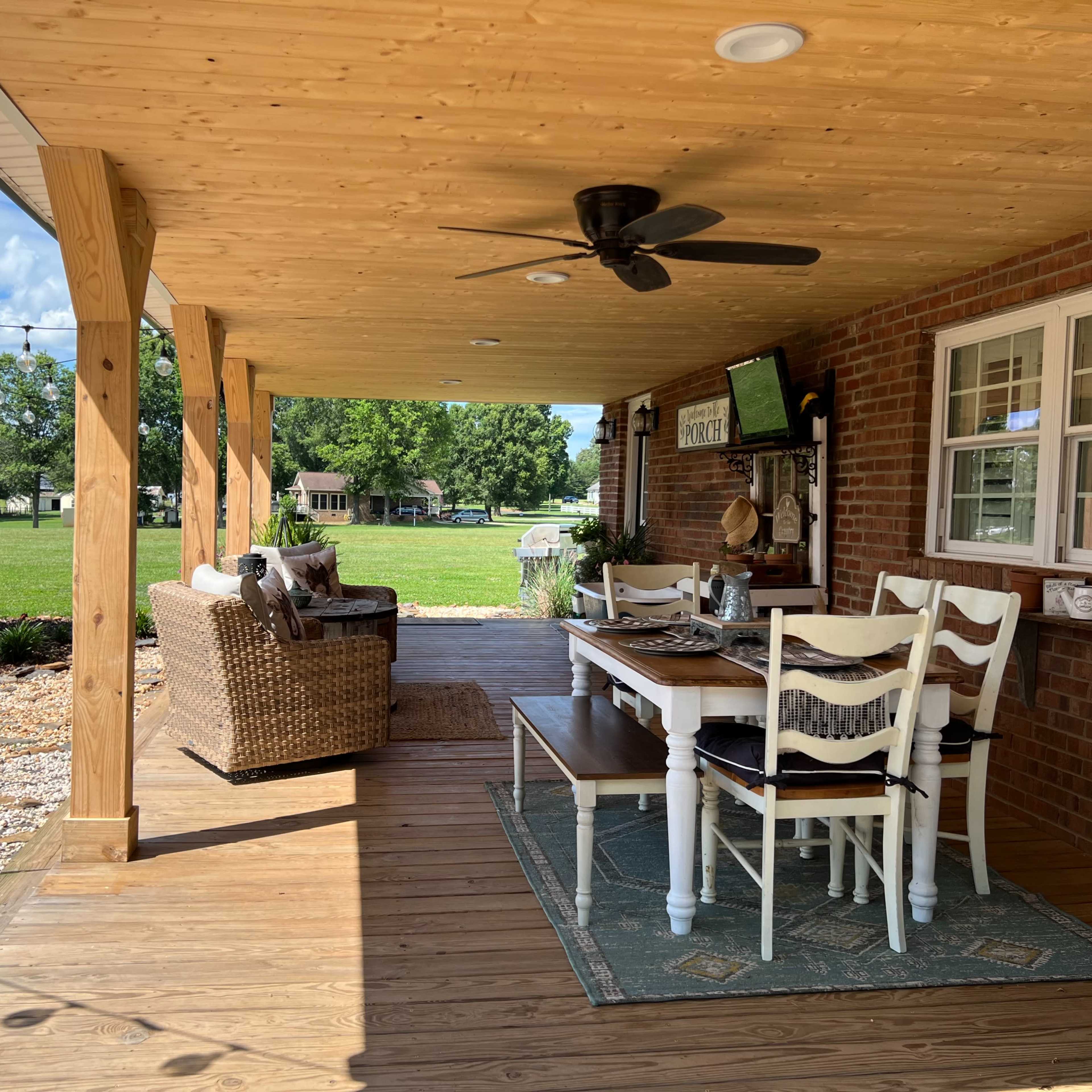 A covered porch features a dining table, chairs, and a seating area, surrounded by green grass and trees.