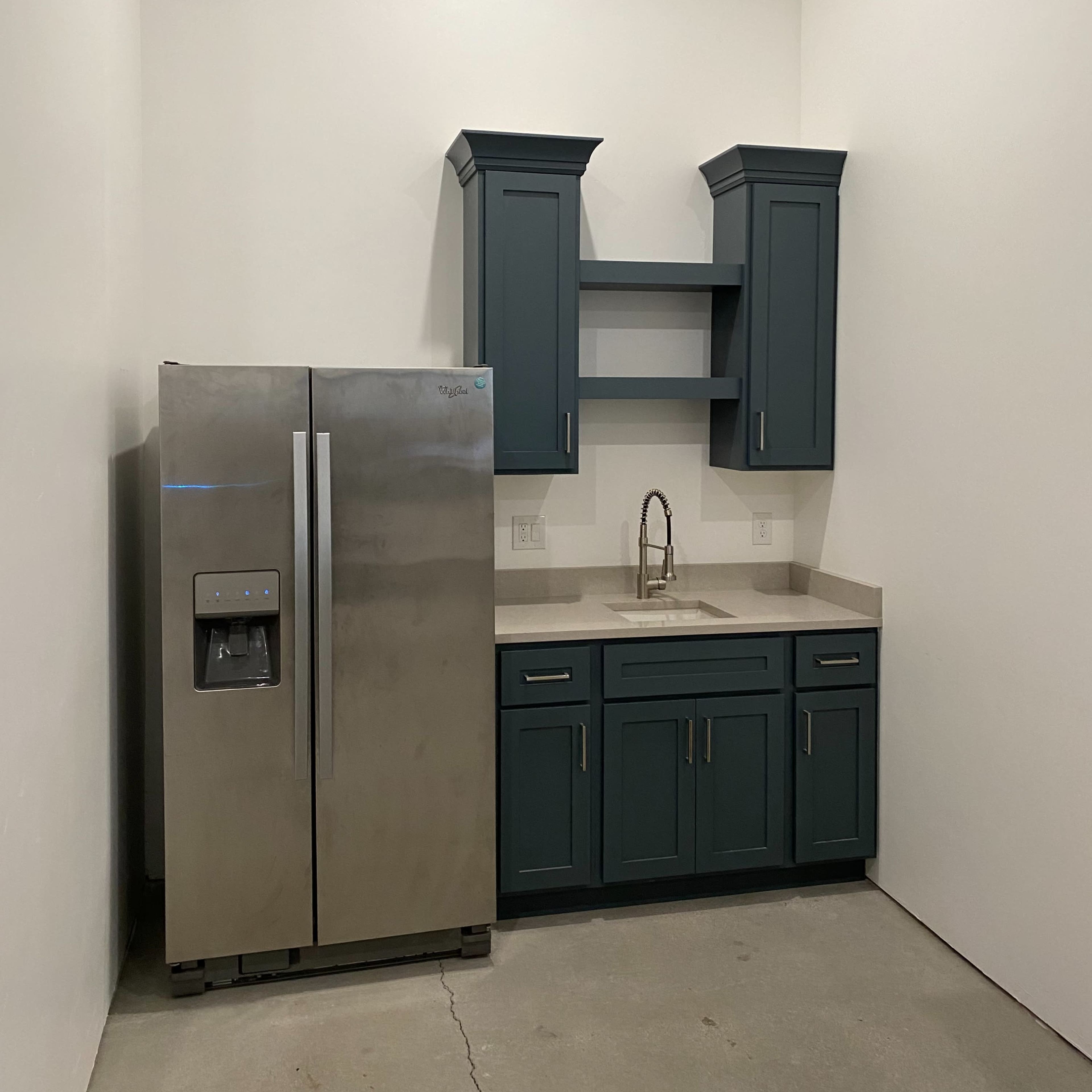 A kitchen corner features a stainless steel refrigerator beside dark blue cabinets and a sink with a countertop.