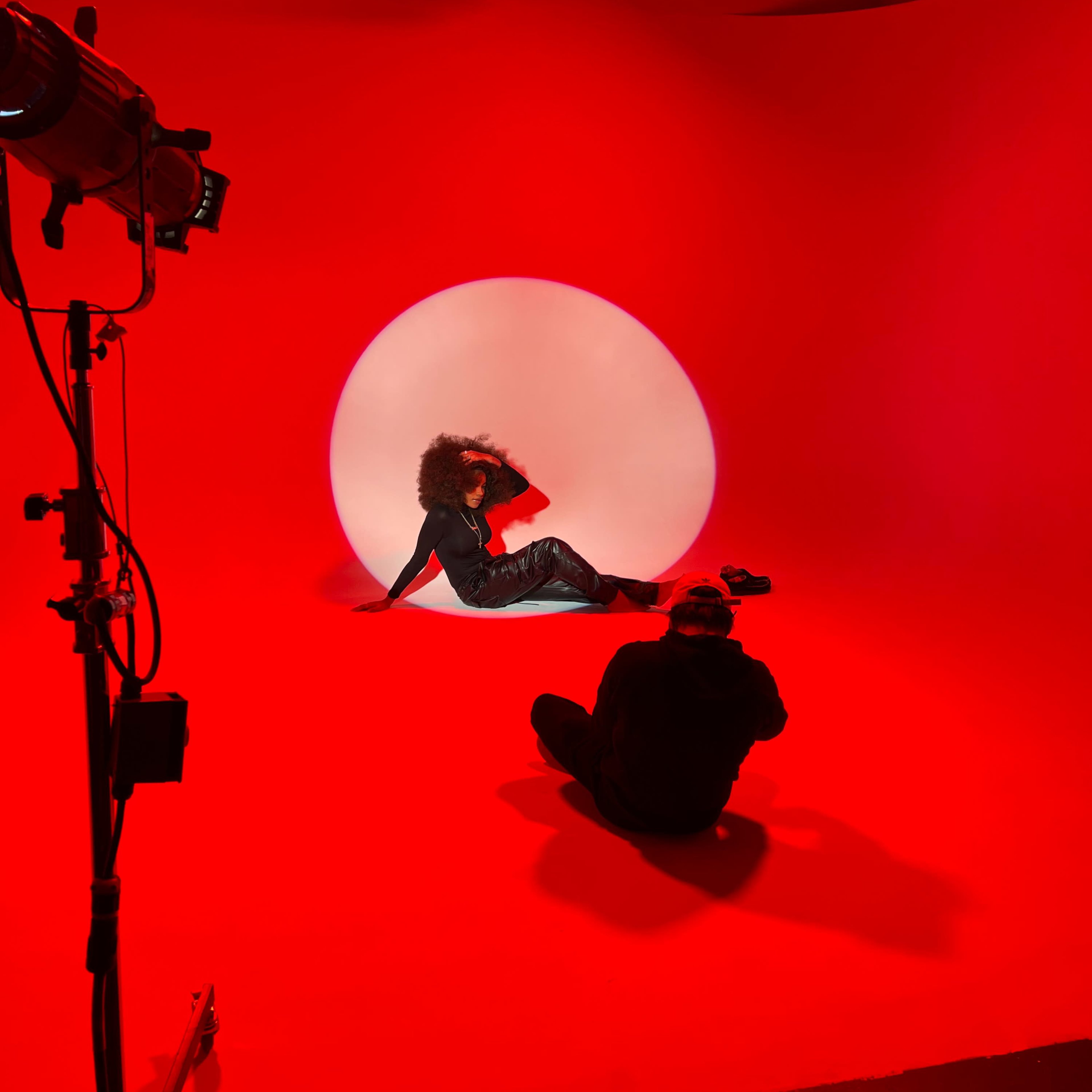 A model poses against a red backdrop with a circular light while a photographer captures the scene.