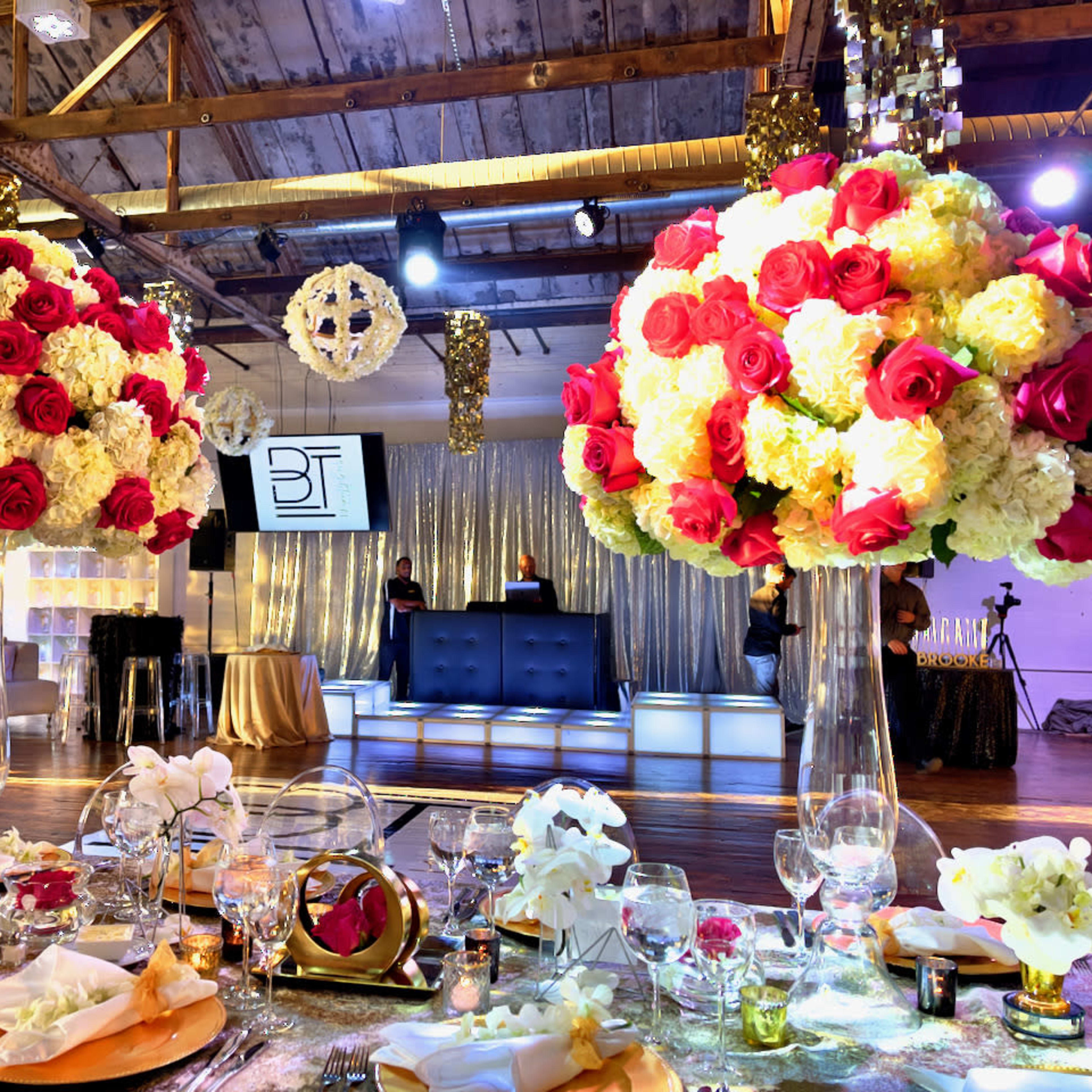 A wedding reception setup with large floral centerpieces of roses and hydrangeas, elegantly decorated tables, and a DJ booth in the background.