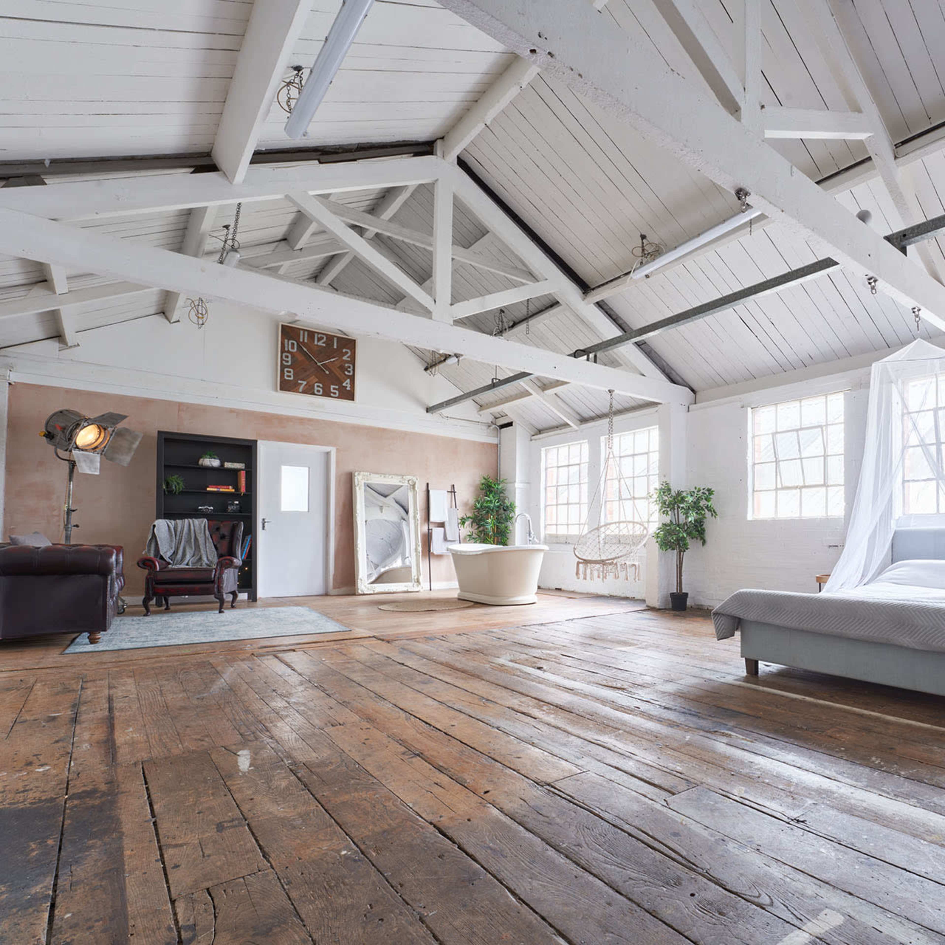 A spacious loft-style bedroom features a bed with a sheer canopy, large windows, and exposed wooden beams, with a freestanding bathtub and a cozy sitting area in the background.