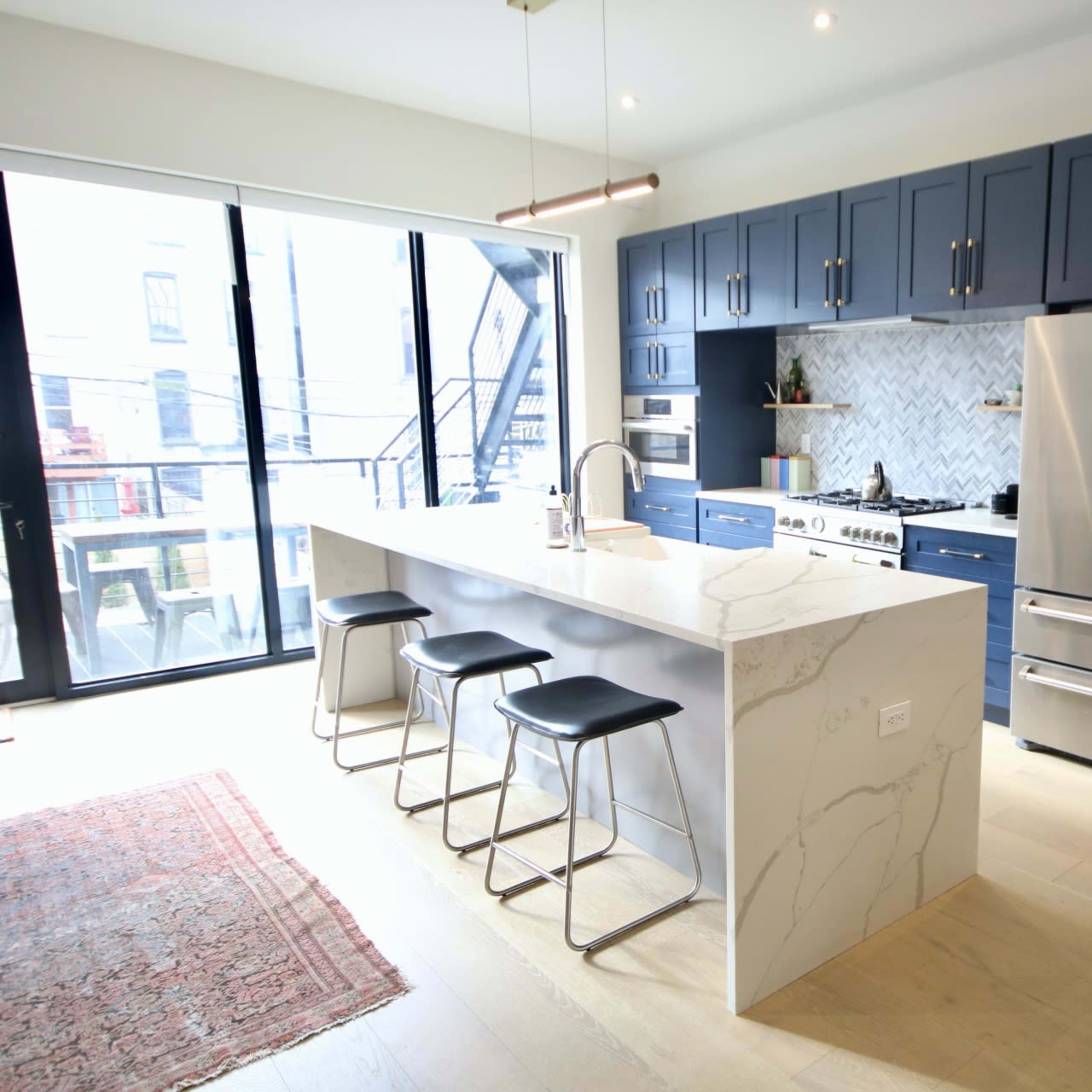 A modern kitchen features a large island with a white marble countertop, dark blue cabinets, and stainless steel appliances.