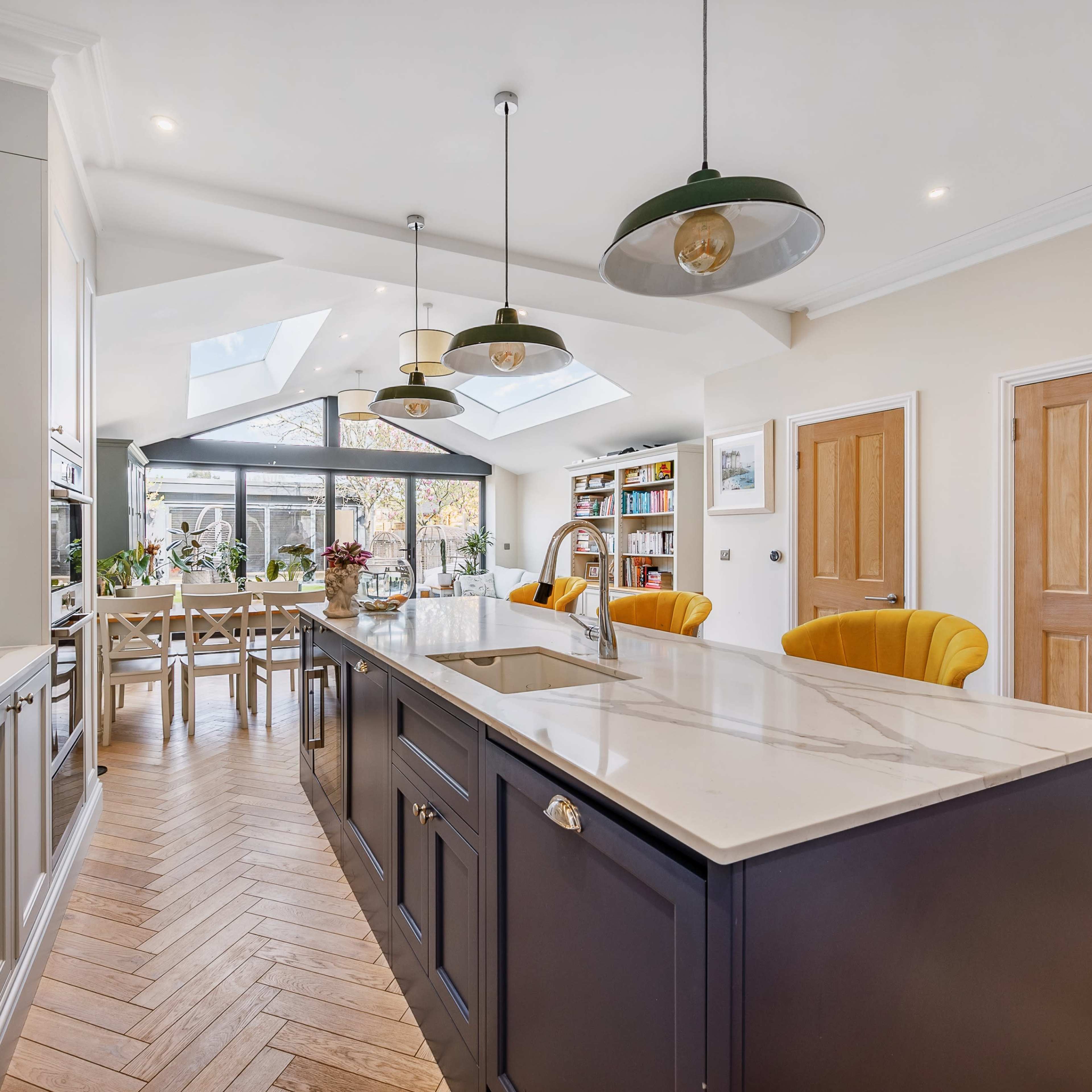 A modern kitchen features a central island with bar stools, sleek cabinetry, and large windows overlooking a backyard patio.