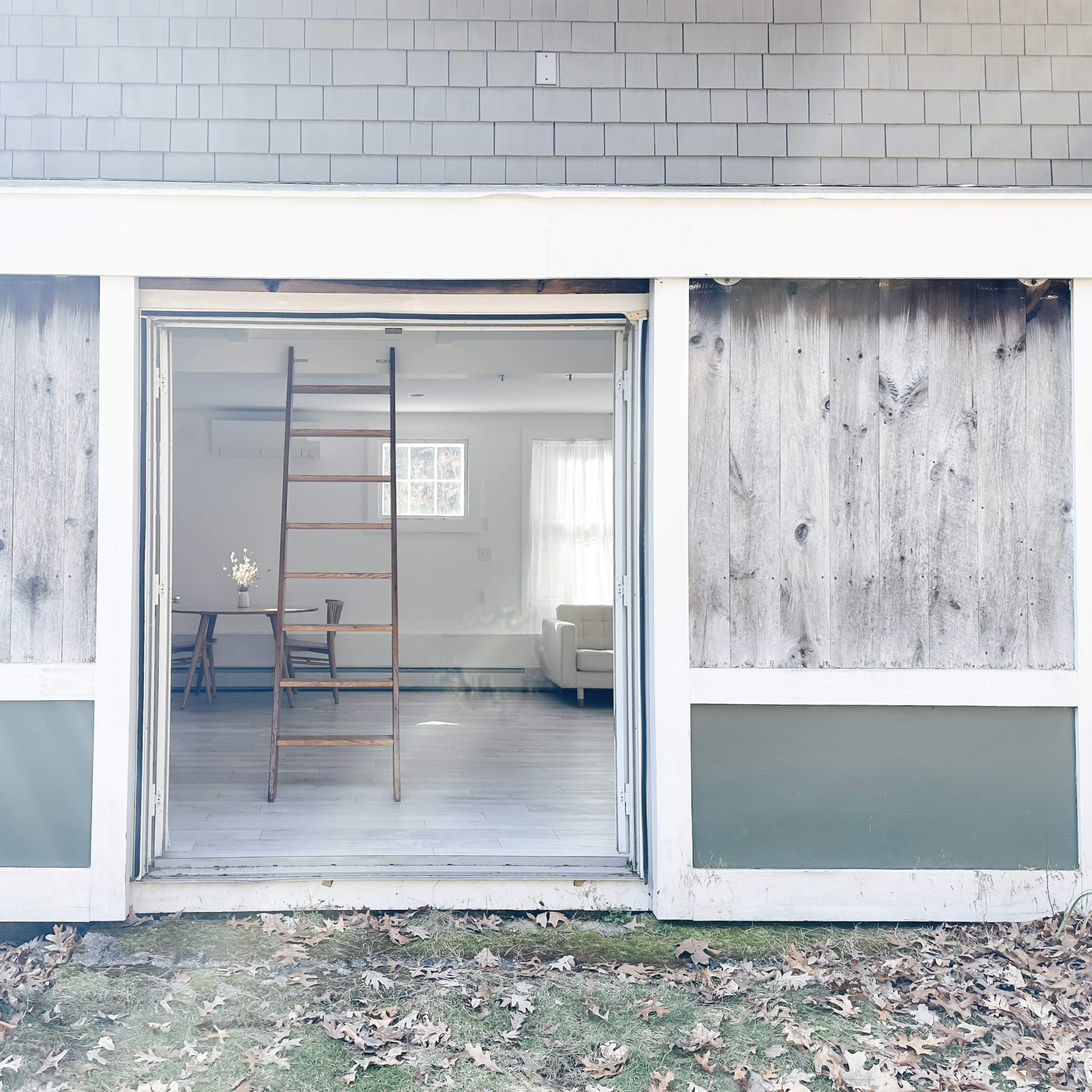 A wooden ladder leans against an open doorway leading into a room with a dining table and chairs.