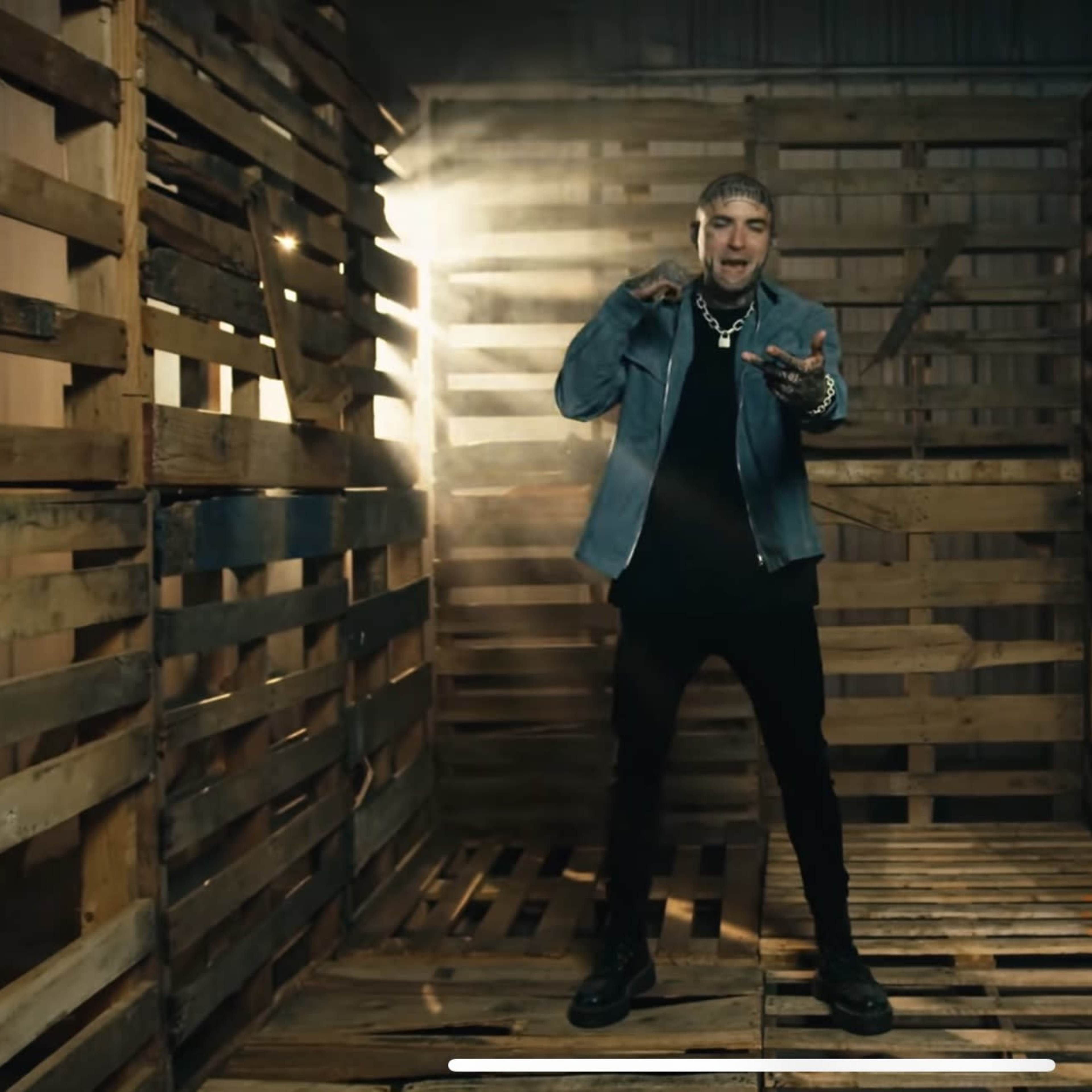 A person stands in a dimly lit wooden structure made of pallets, holding a microphone in a performance stance.
