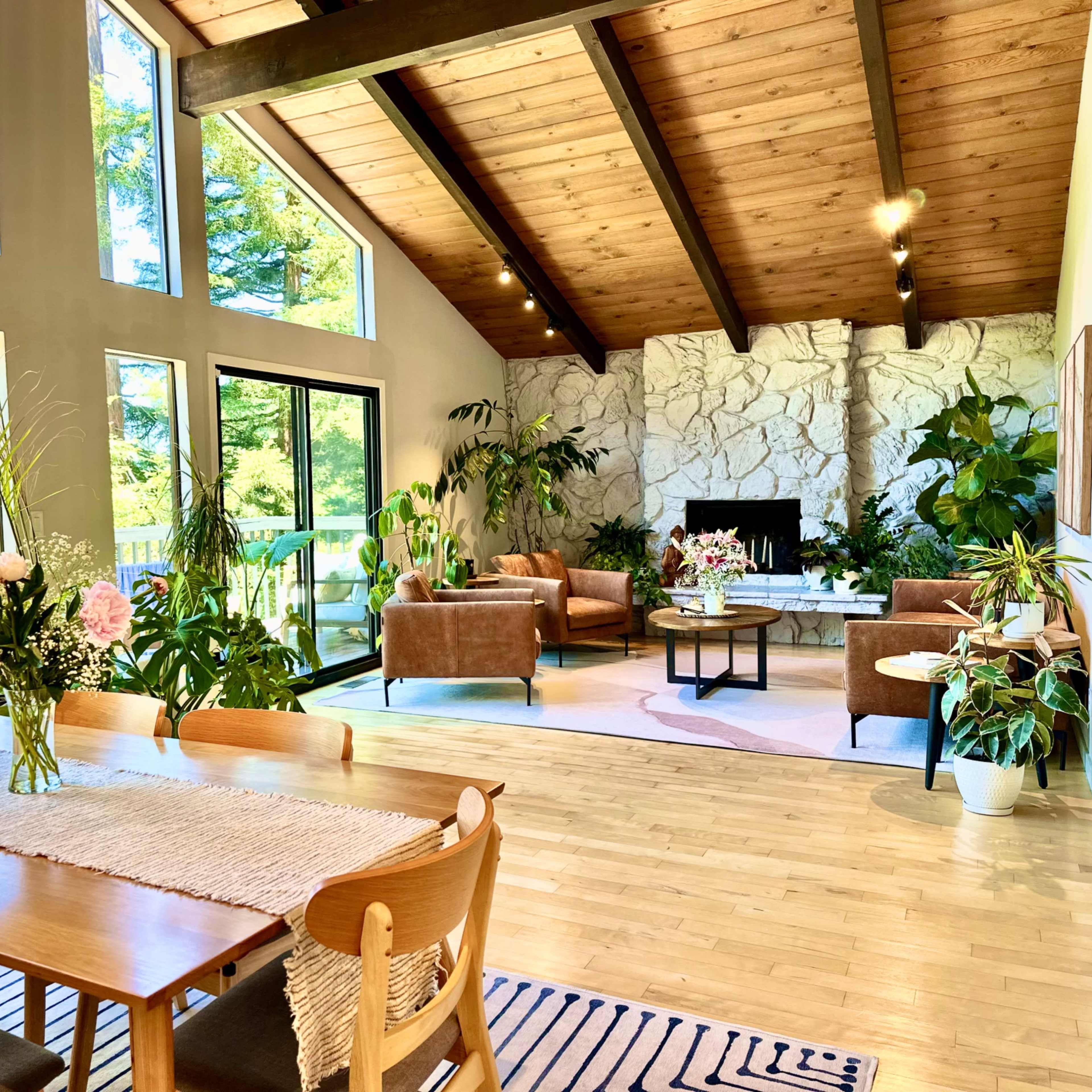 A spacious living area features large windows, wooden beams, and a blend of modern and natural decor, including a dining table and comfortable seating surrounded by greenery.