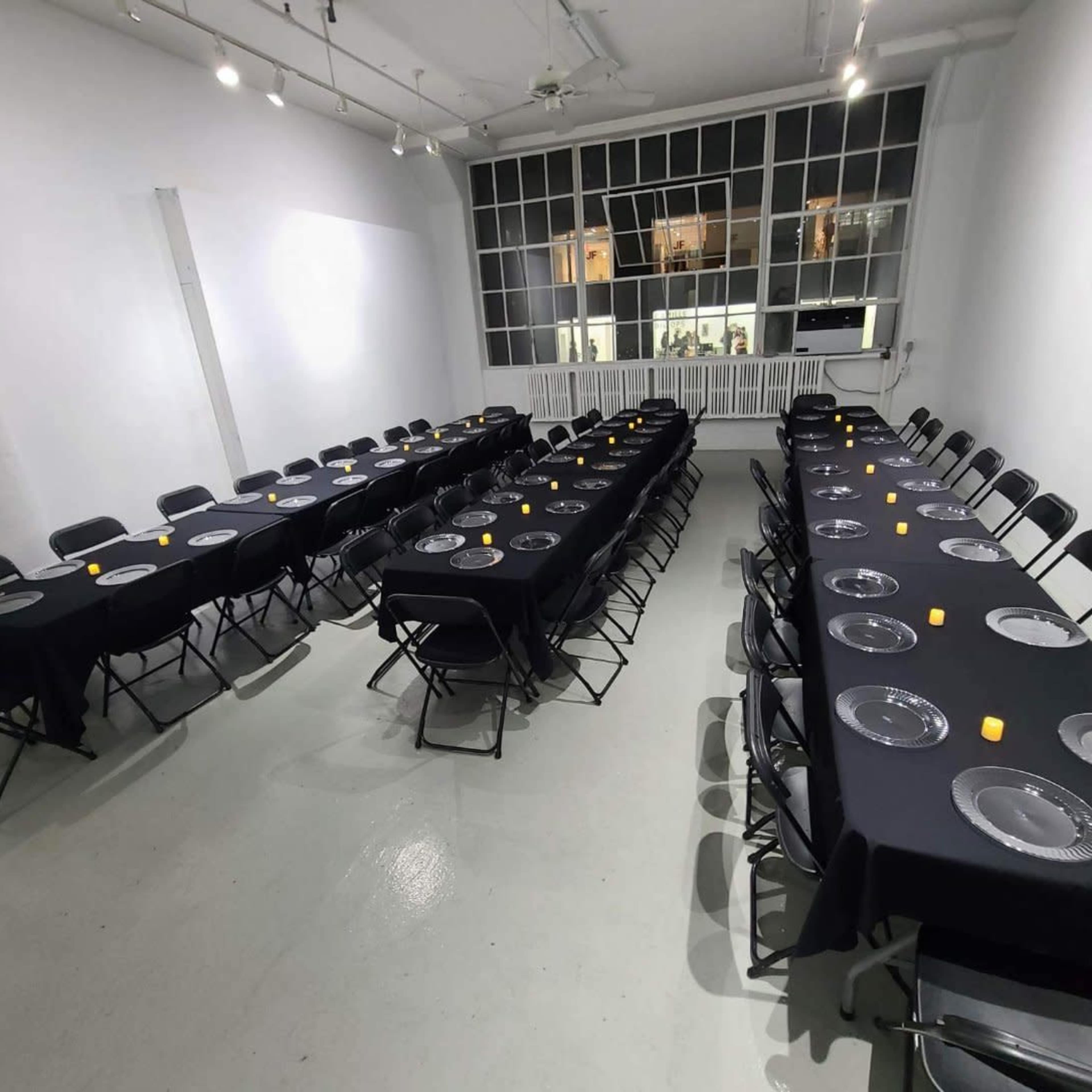 A long, rectangular table is set up in a bright, white room, covered with black tablecloths and arranged with clear plates and small yellow candles.