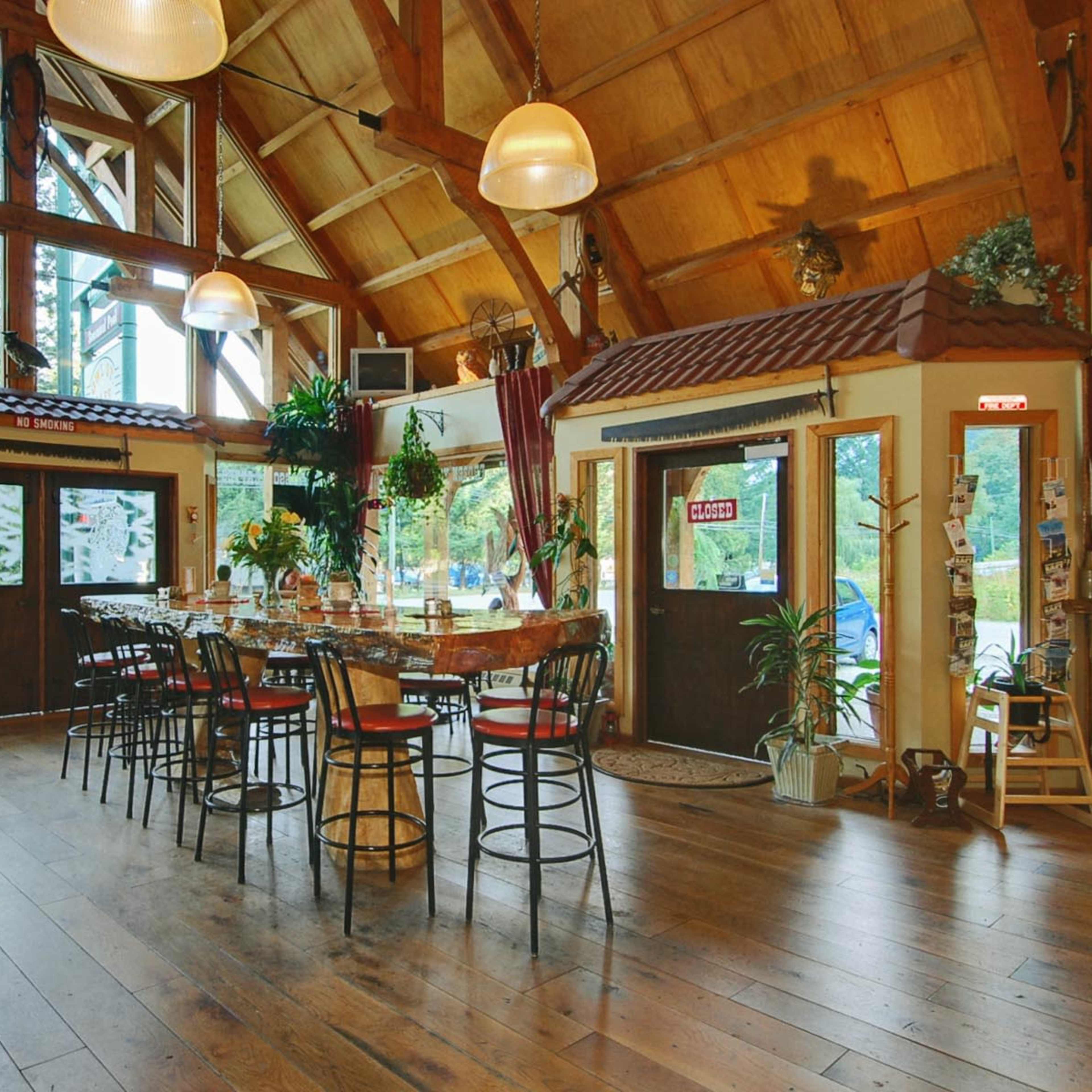 The interior of a rustic dining establishment features wooden beams, large windows, and a central communal table surrounded by high chairs.
