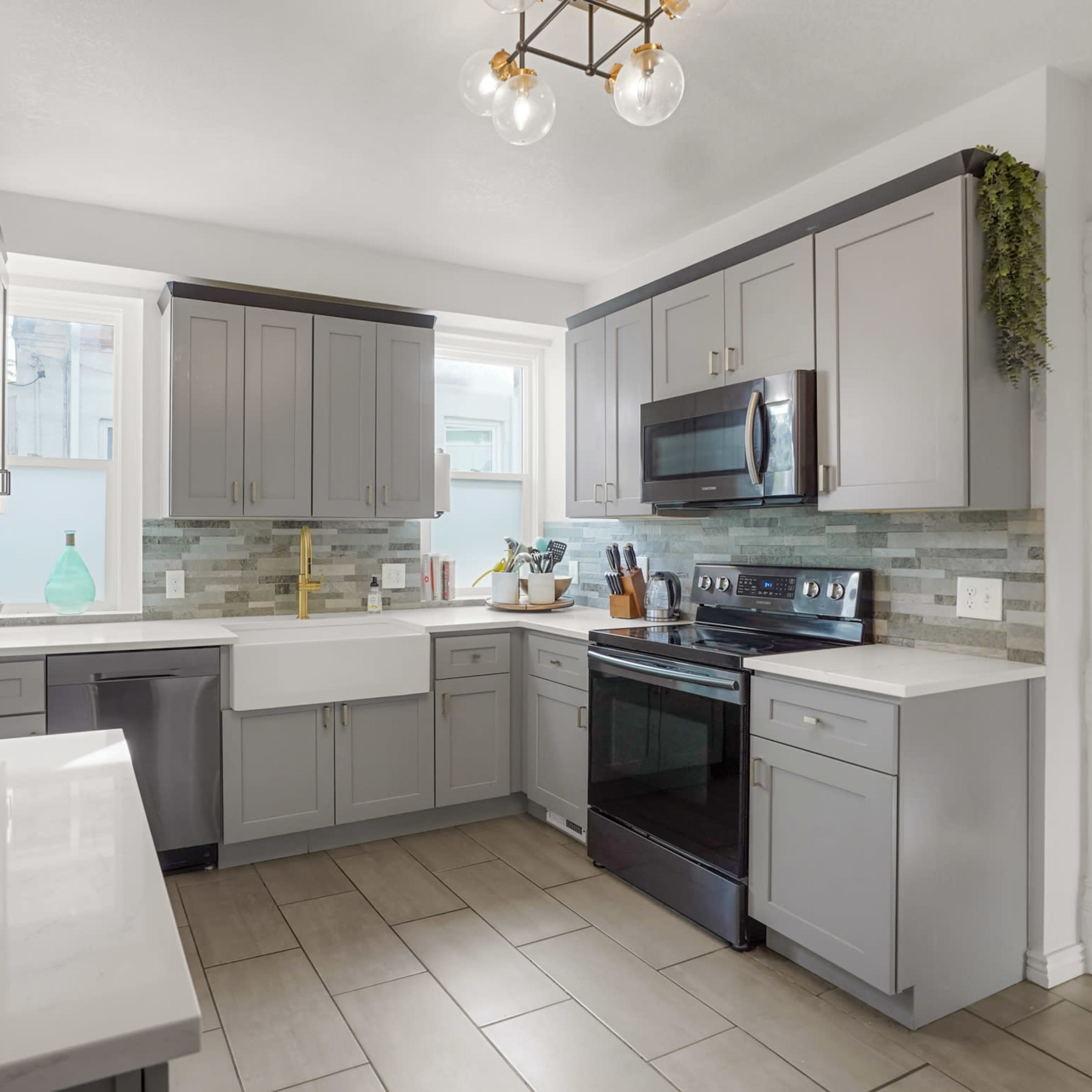A modern kitchen featuring gray cabinetry, a farmhouse sink, stainless steel appliances, and neutral tile flooring.