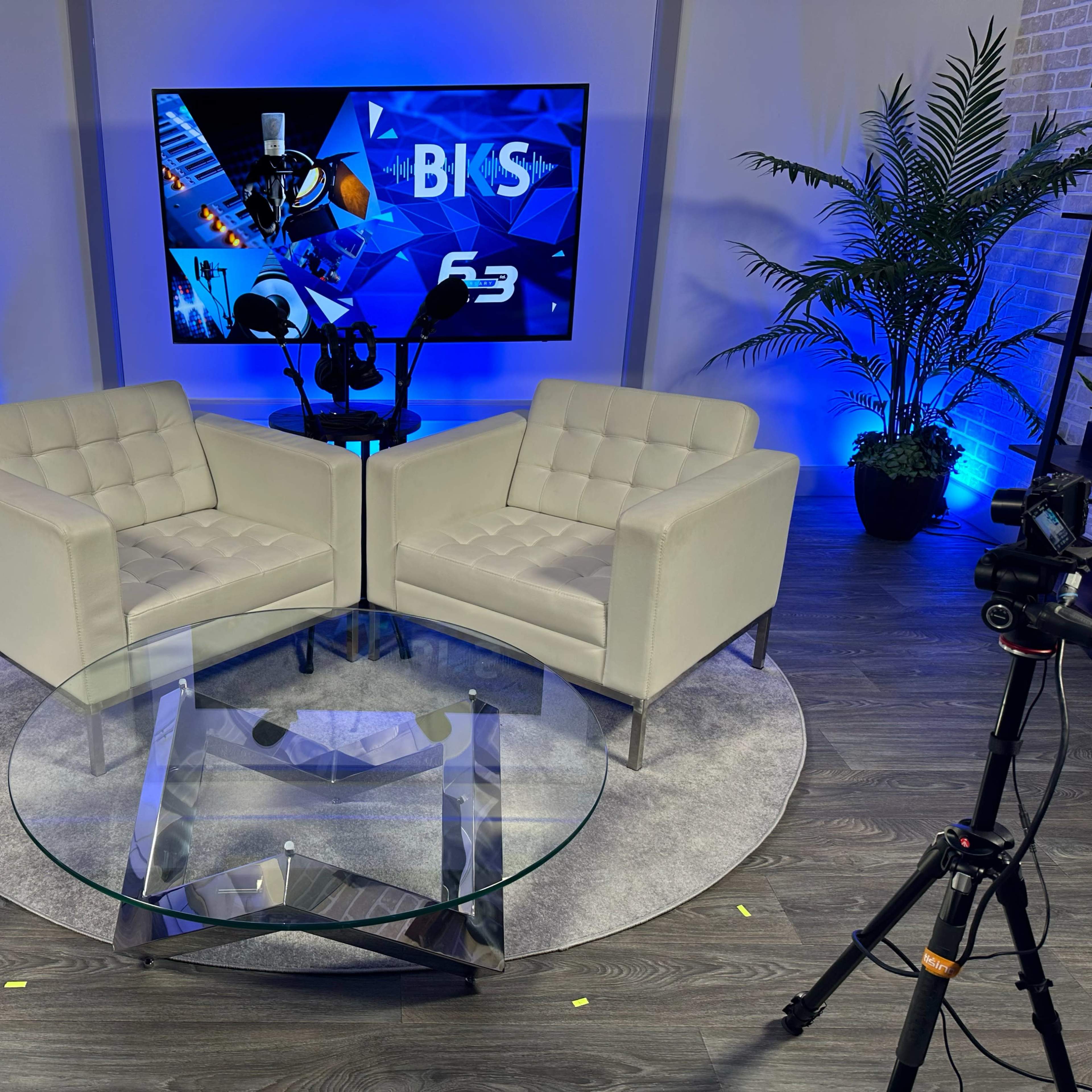 A modern interview setup with two white armchairs facing each other around a glass coffee table, framed by a blue-lit backdrop featuring a screen displaying graphics.