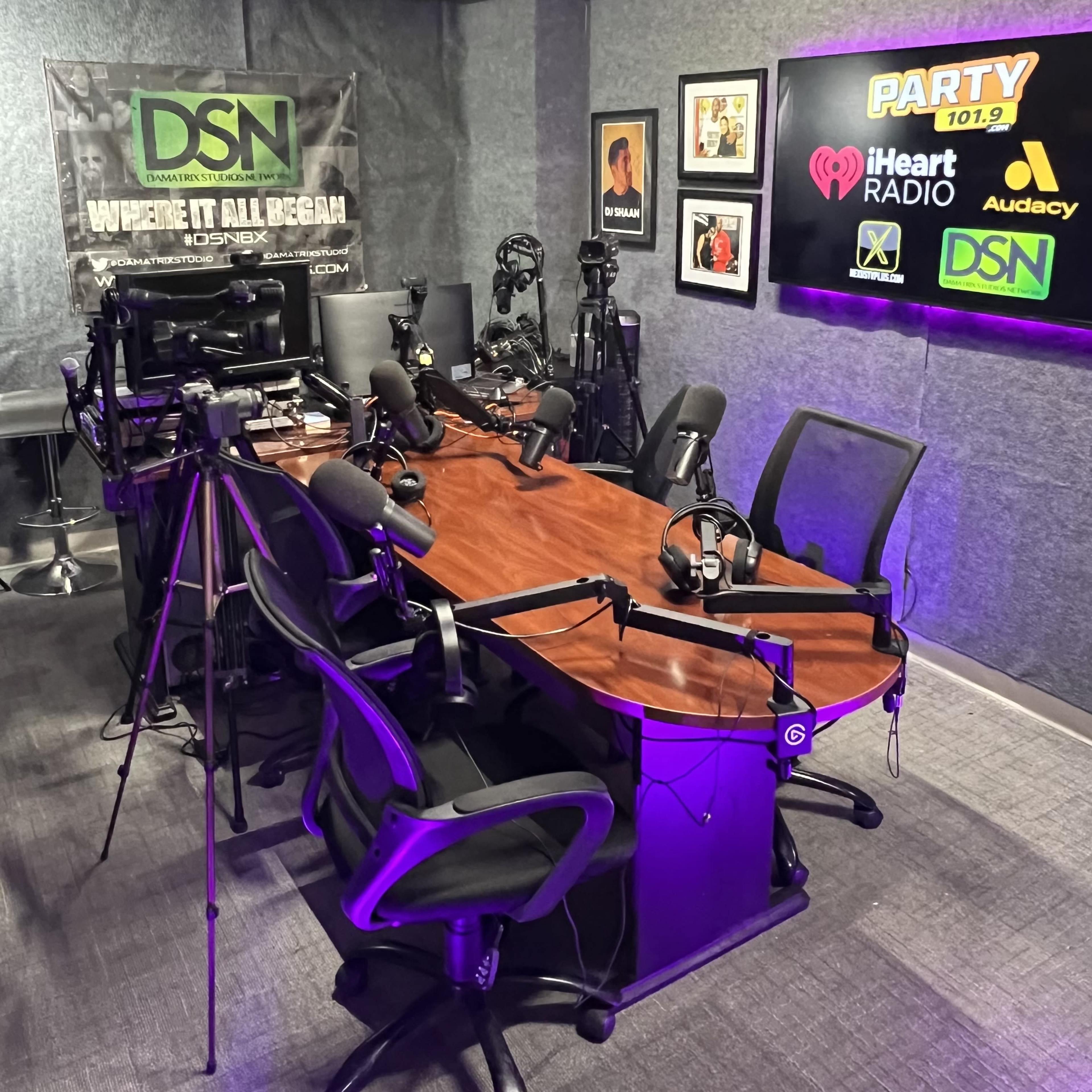 A radio studio setup with a large table, several microphones, chairs, and a wall displaying logos and screens for various media platforms.