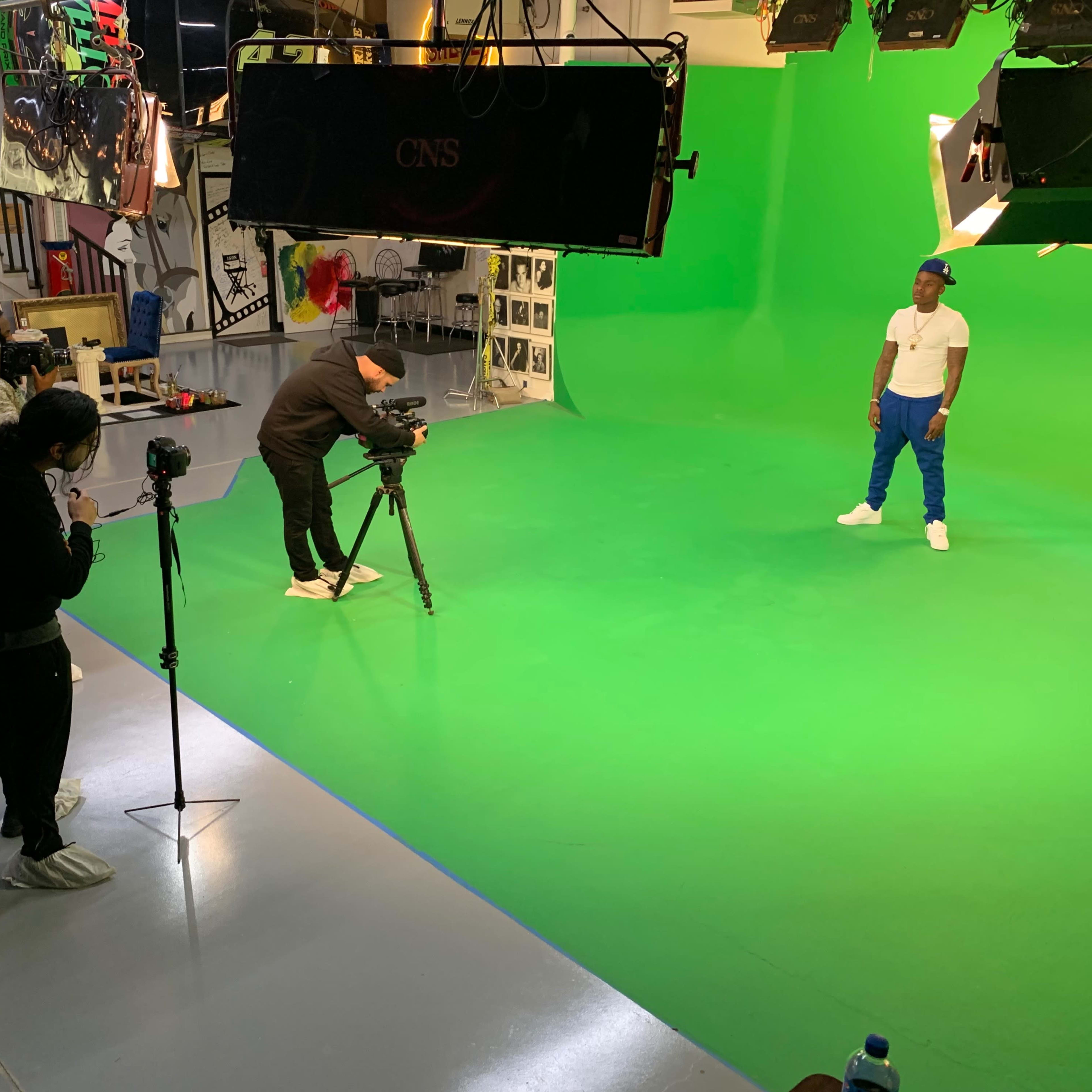 A film crew sets up equipment and prepares to shoot a scene with an actor on a green screen backdrop.