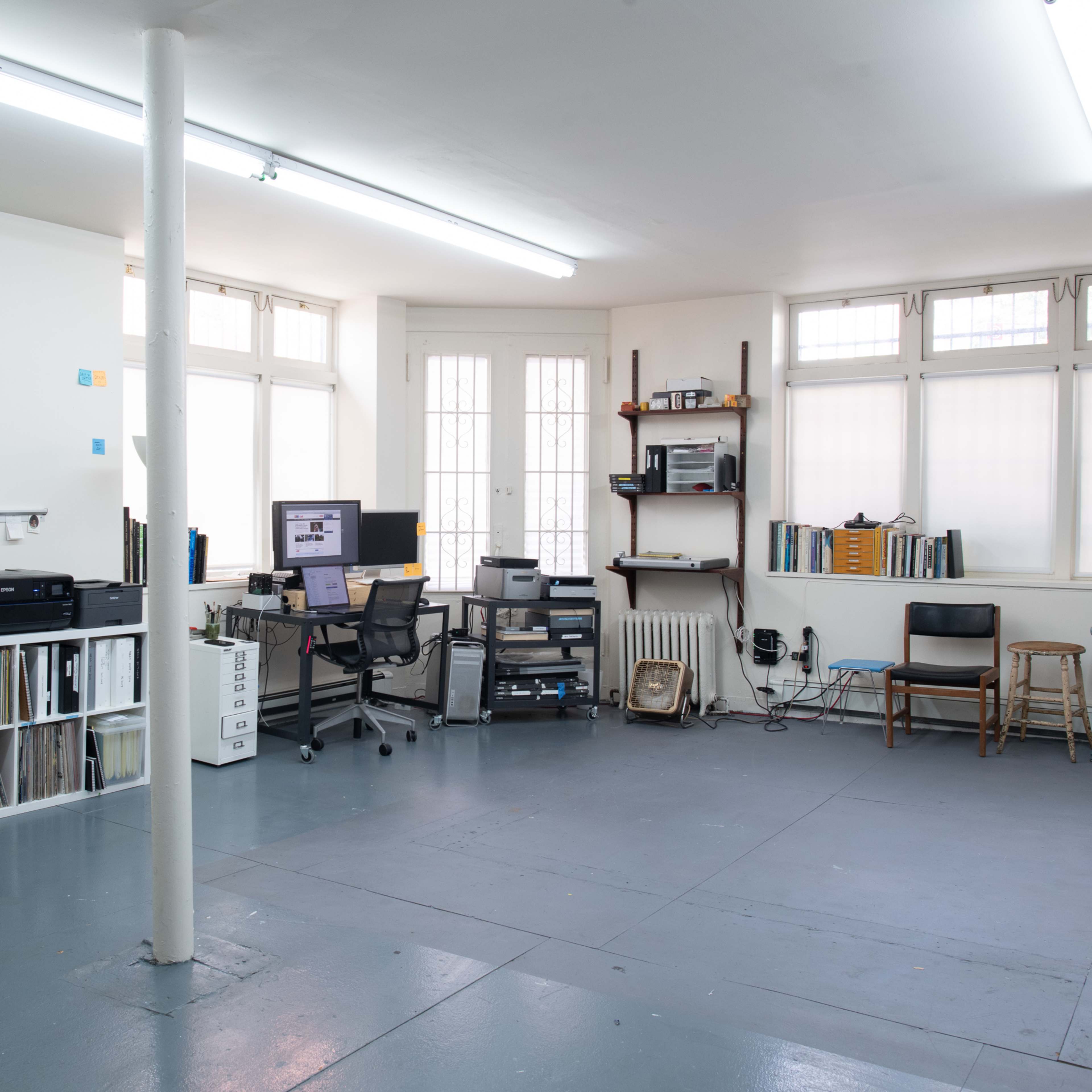 A spacious, minimally furnished office or studio with a desk, computer setup, shelves, and a printer against the walls, featuring large windows that provide natural light.