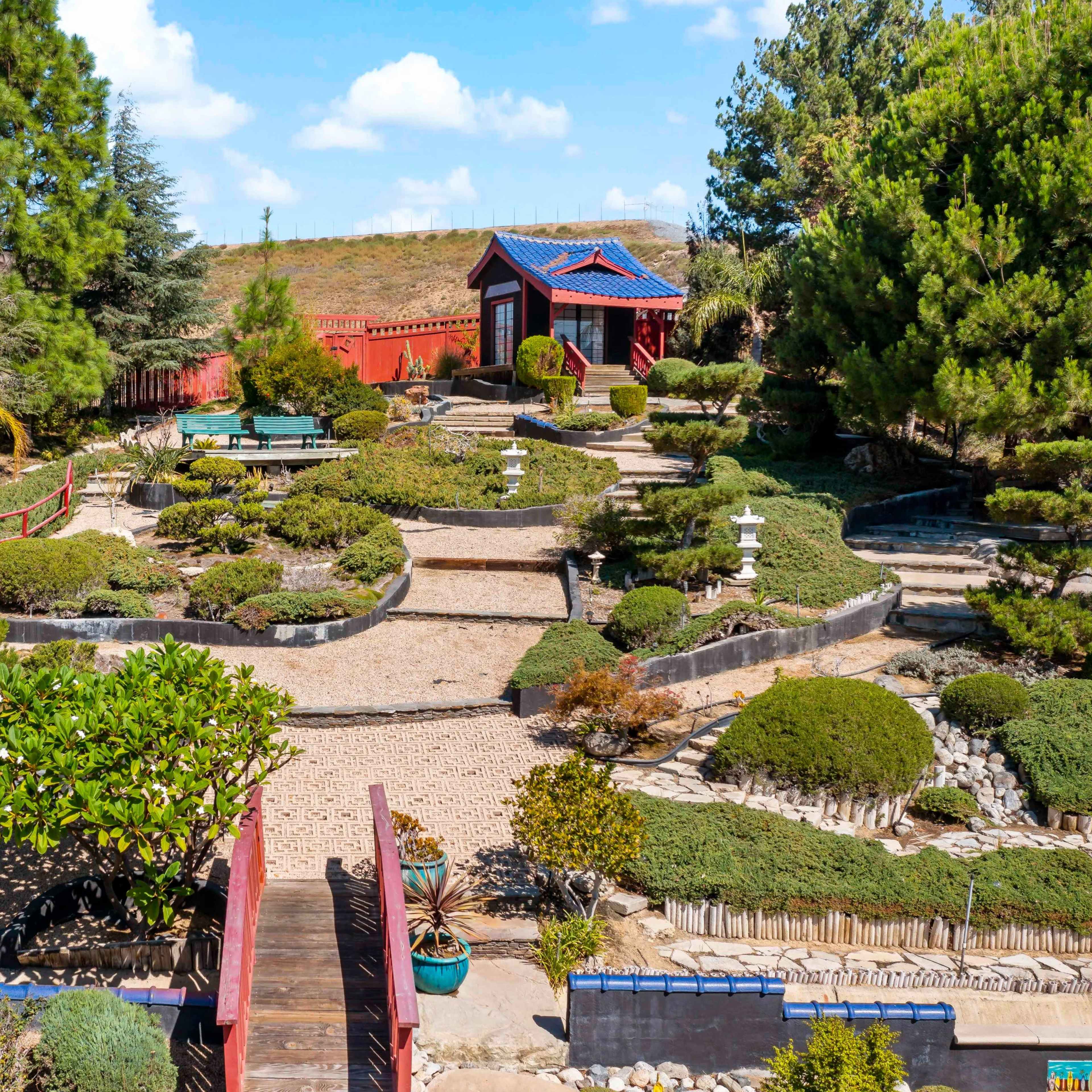 A landscaped garden featuring winding paths, manicured shrubs, a pond, and a pagoda-style building surrounded by trees.