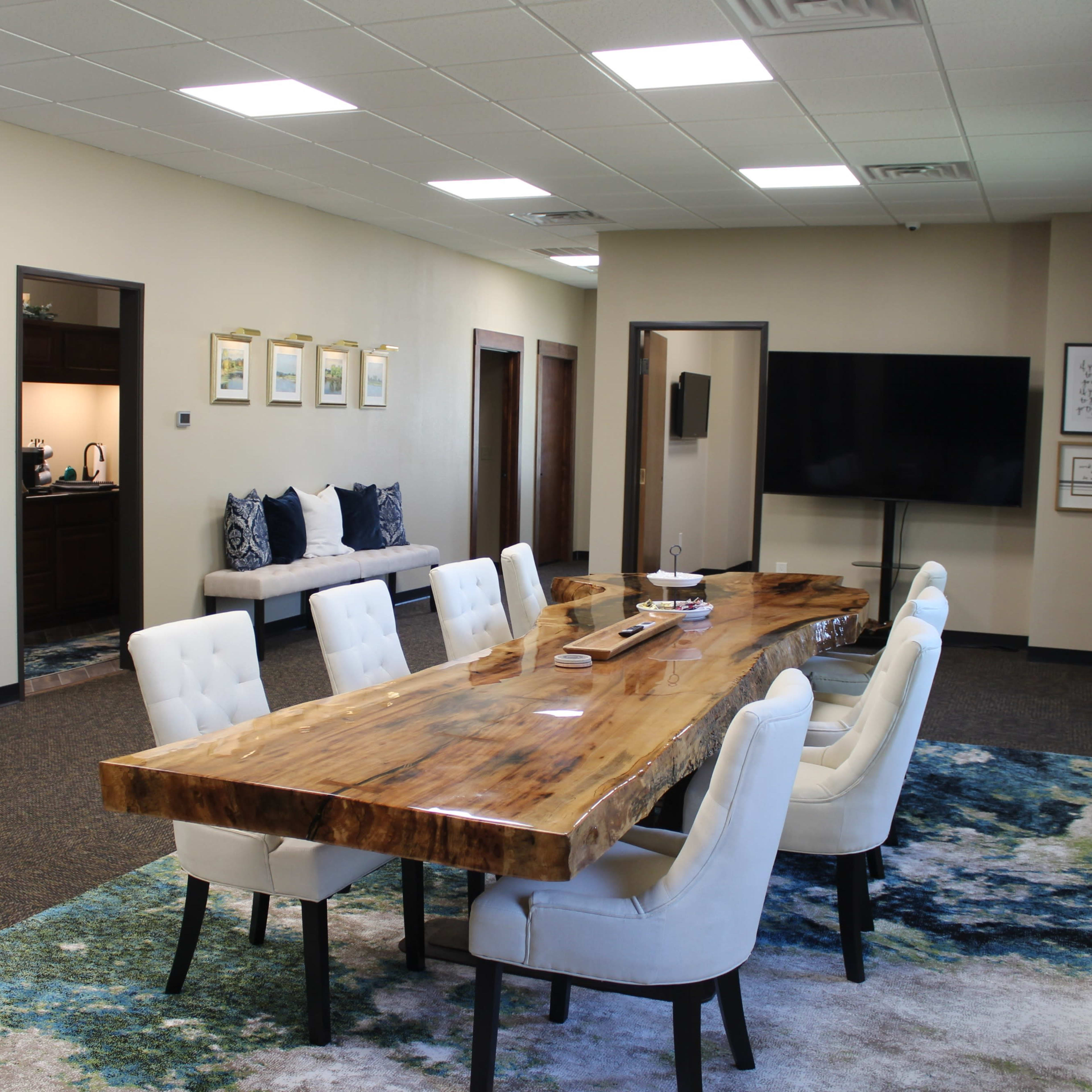 A spacious meeting room with a large wooden table surrounded by white chairs, a television mounted on the wall, and a cozy seating area in the background.
