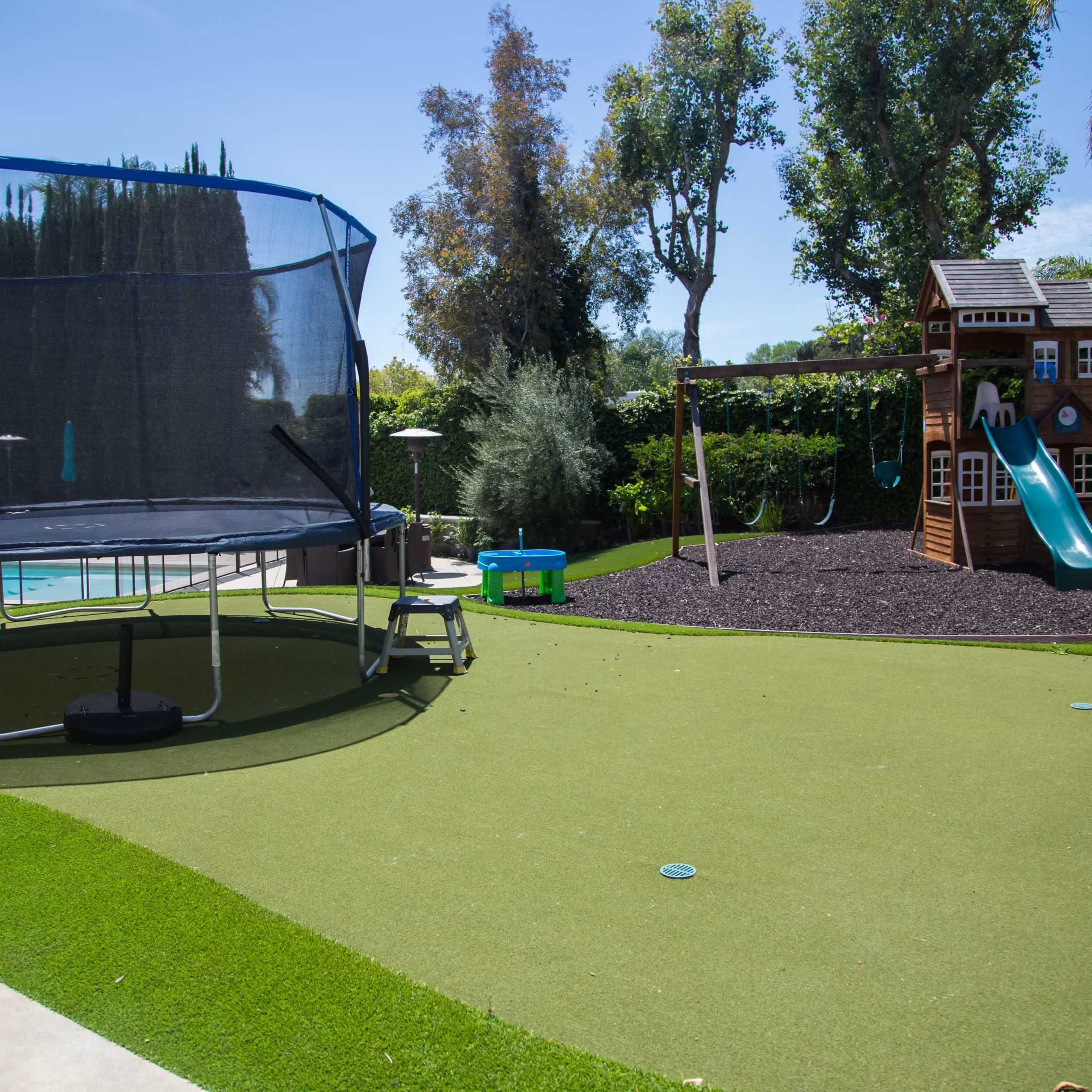 A backyard features a trampoline, a playground structure with a slide, and a landscaped area with artificial grass.