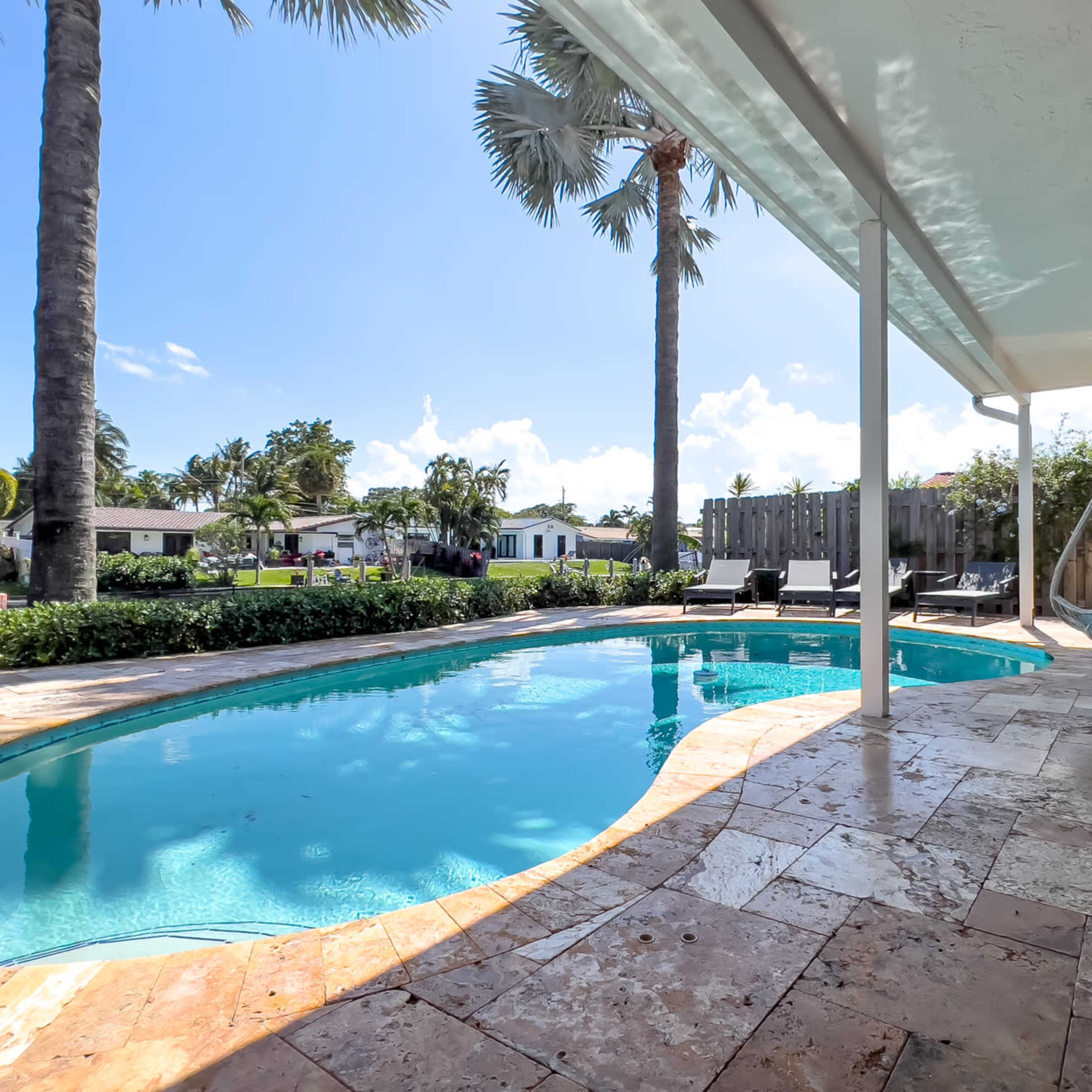 A swimming pool with a hanging chair is surrounded by palm trees and residential yards under a clear blue sky.