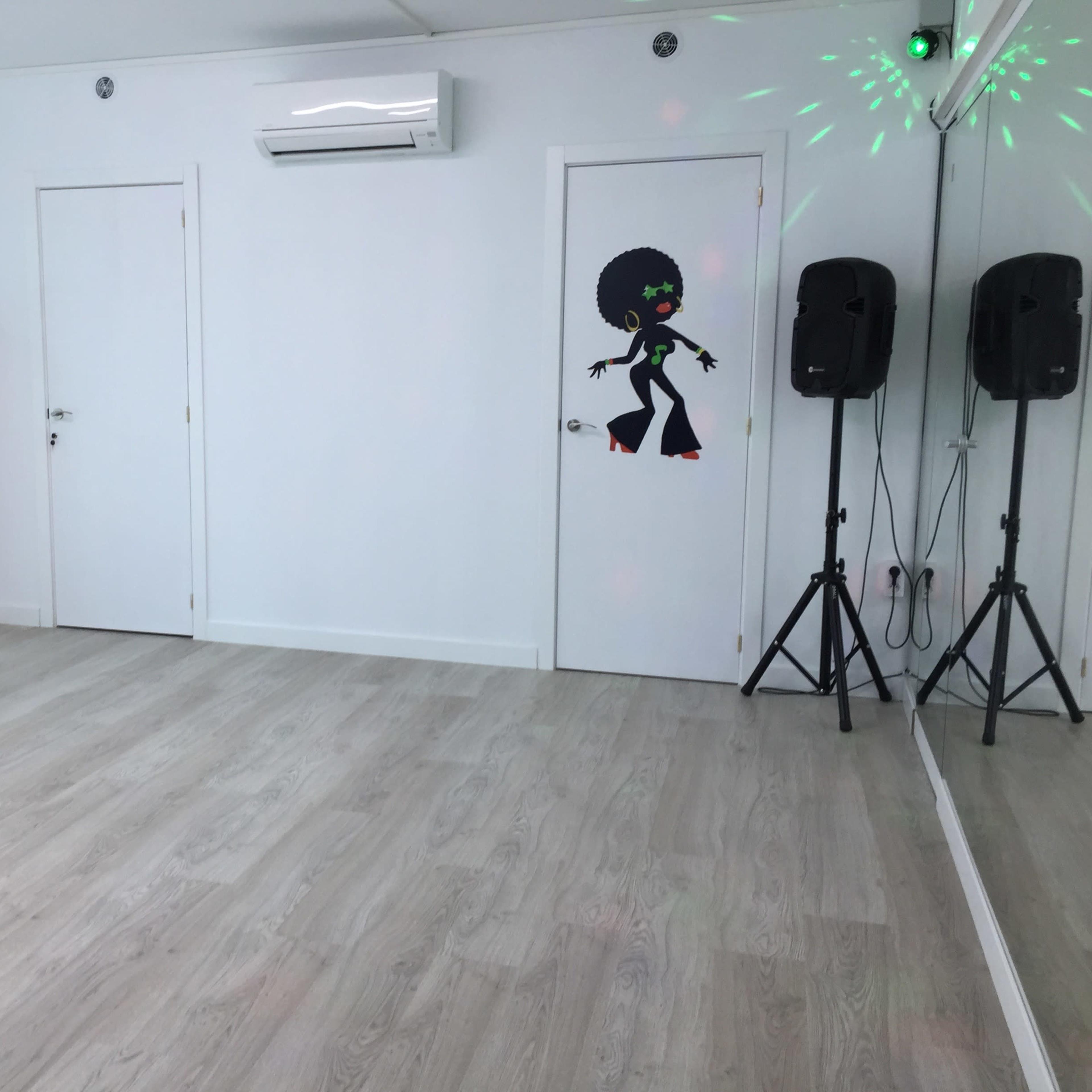 A bright, empty room features a smooth wooden floor, two closed doors, a white shelving unit, and speakers on either side.