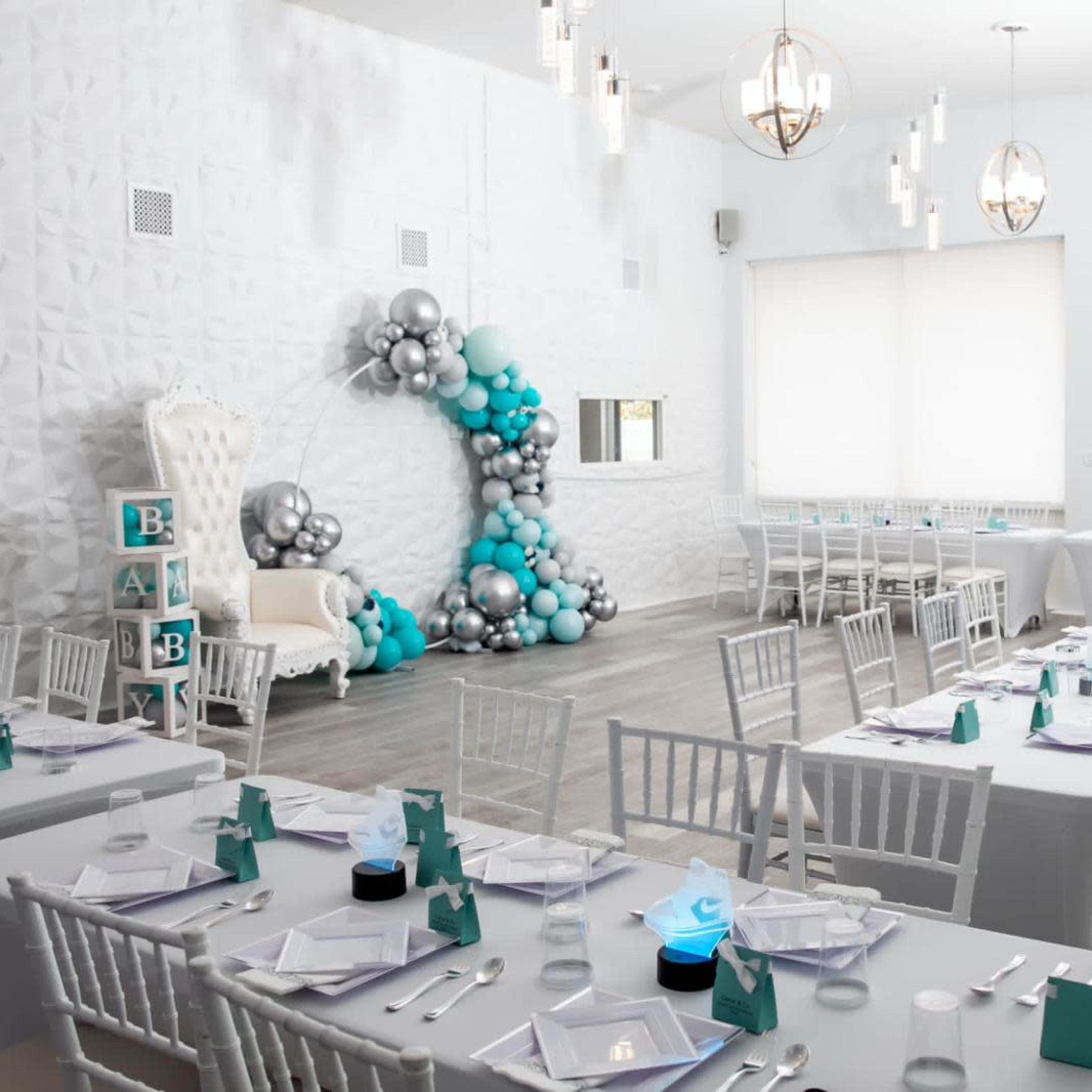 A bright event space arranged with white tables and chairs, featuring a decorative balloon arch and a throne-like chair at one end.