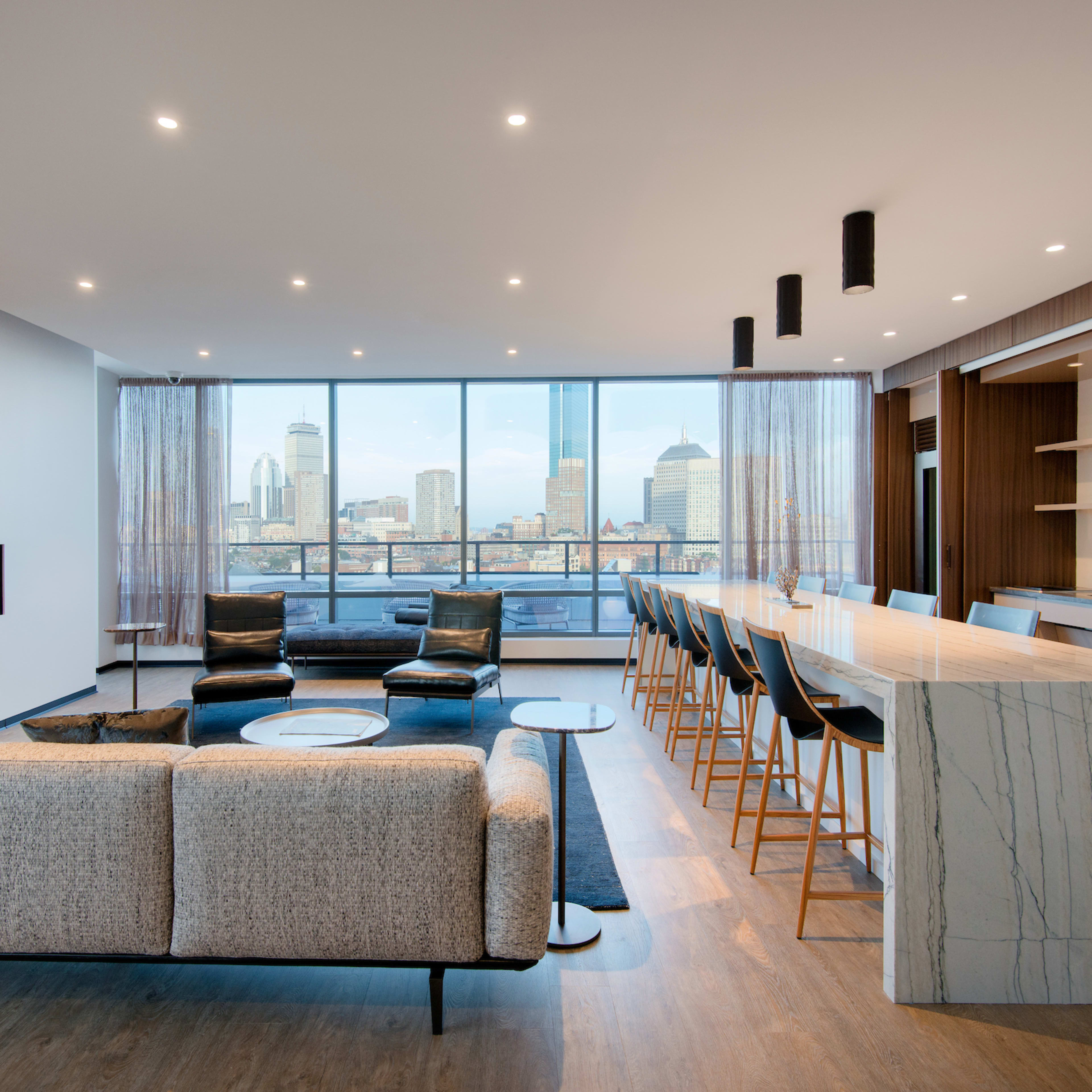 A modern living room features a sectional sofa and a bar-style seating area, with large windows offering a cityscape view and a linear fireplace along one wall.