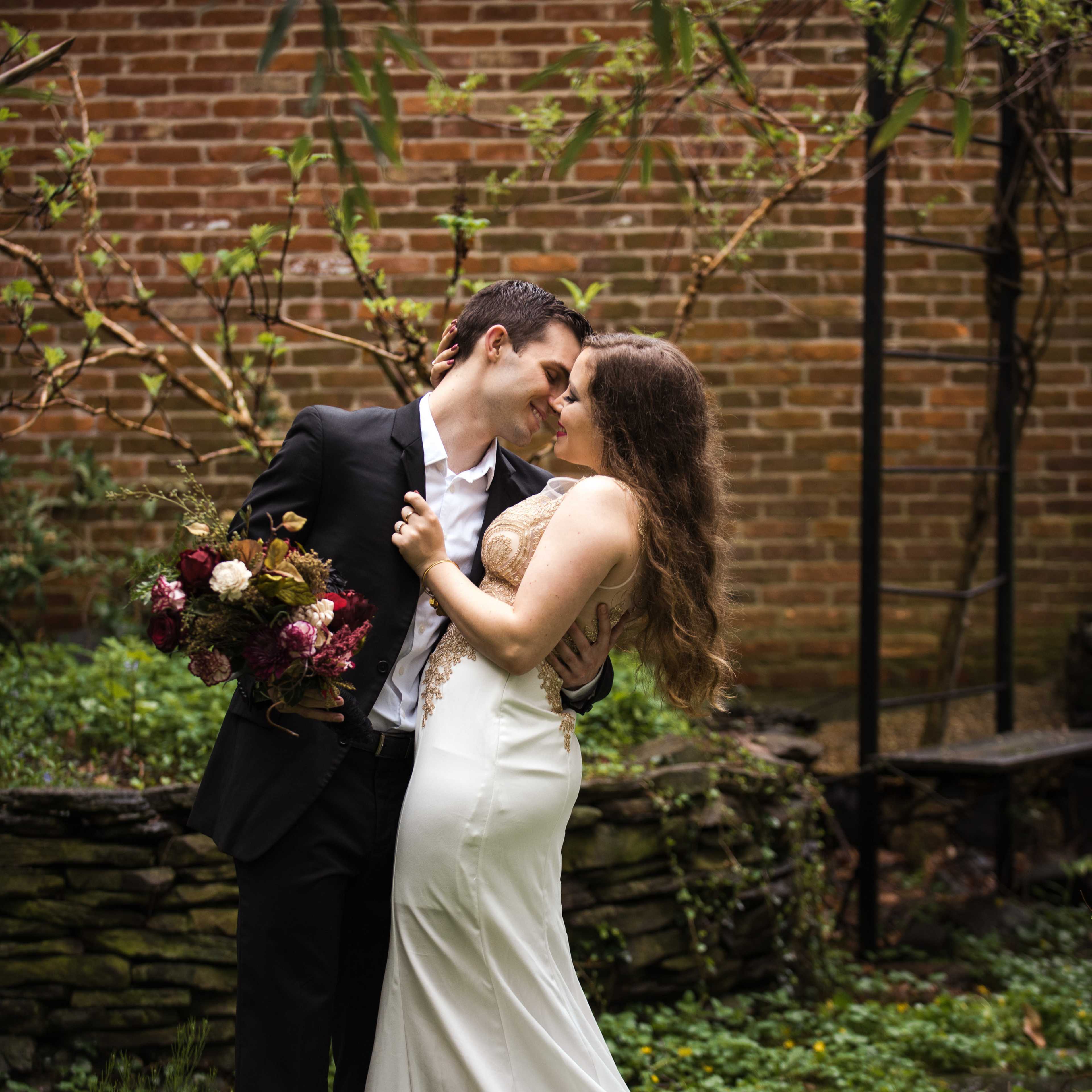 A couple in formal attire embraces in a garden while holding a bouquet, with a brick wall and greenery in the background.