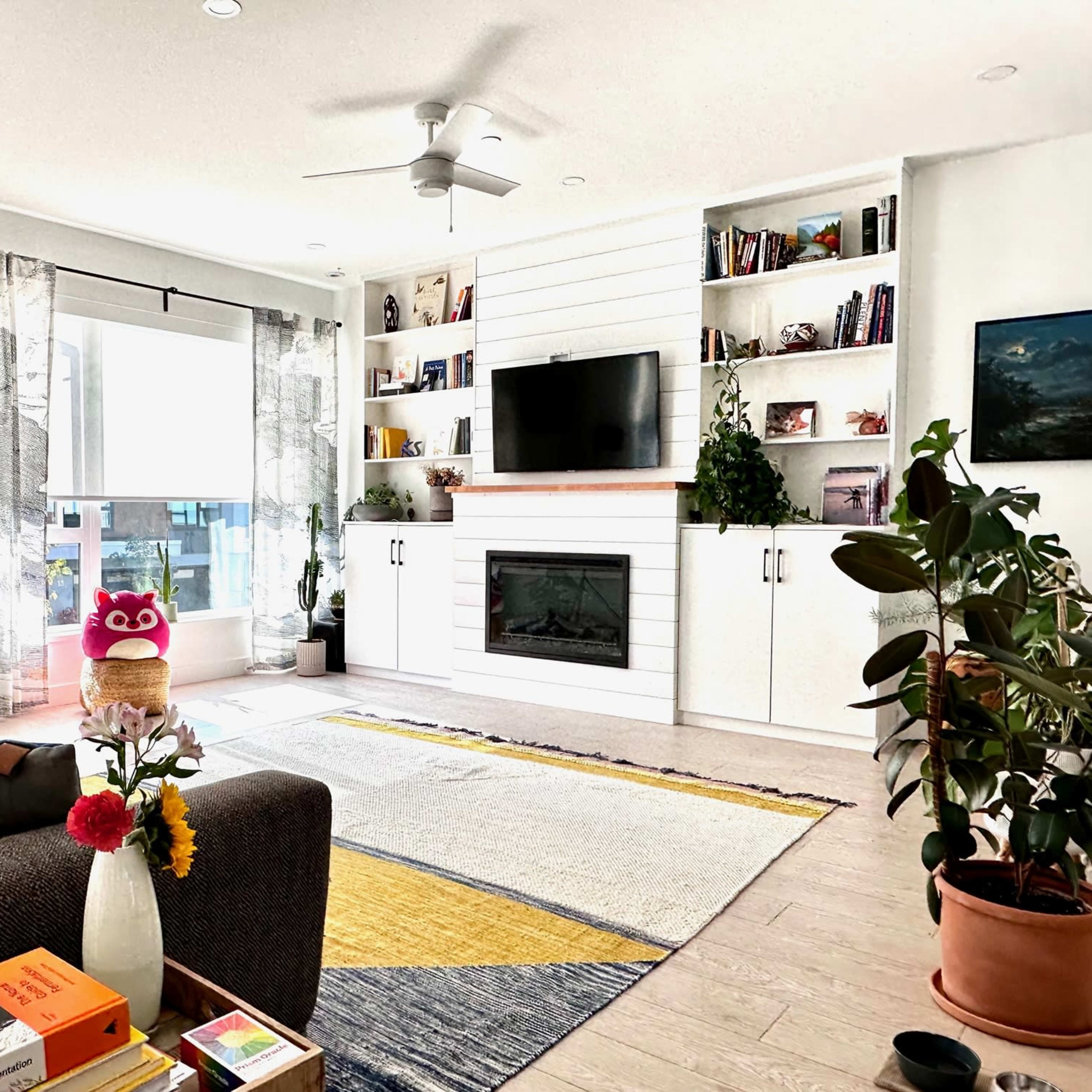 A modern living room featuring a gray sofa, a yellow and gray rug, plants, a white built-in shelf with books and decorative items, and a television above a fireplace.