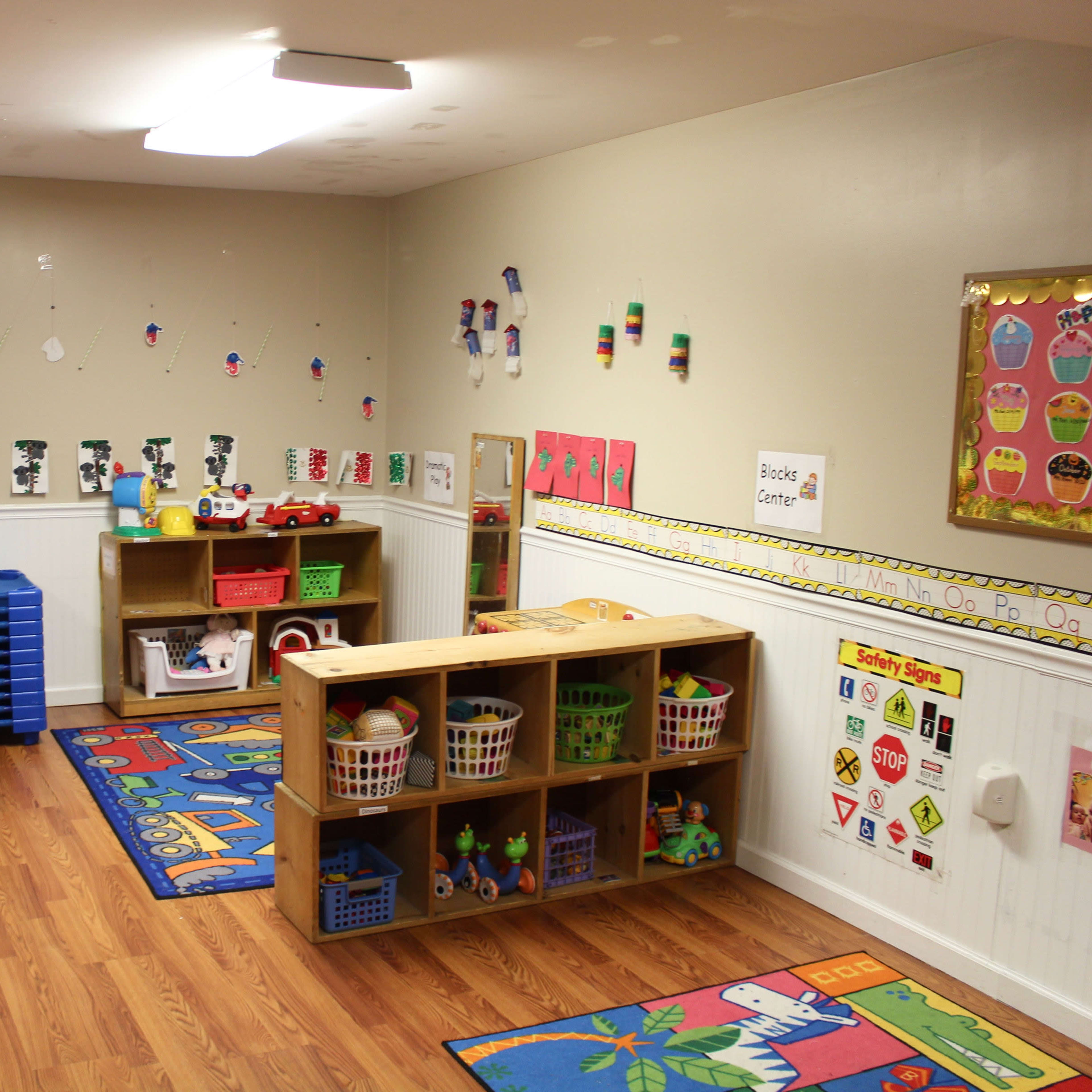 A children's classroom with colorful rugs, storage shelves filled with toys, and labeled areas for different activities.