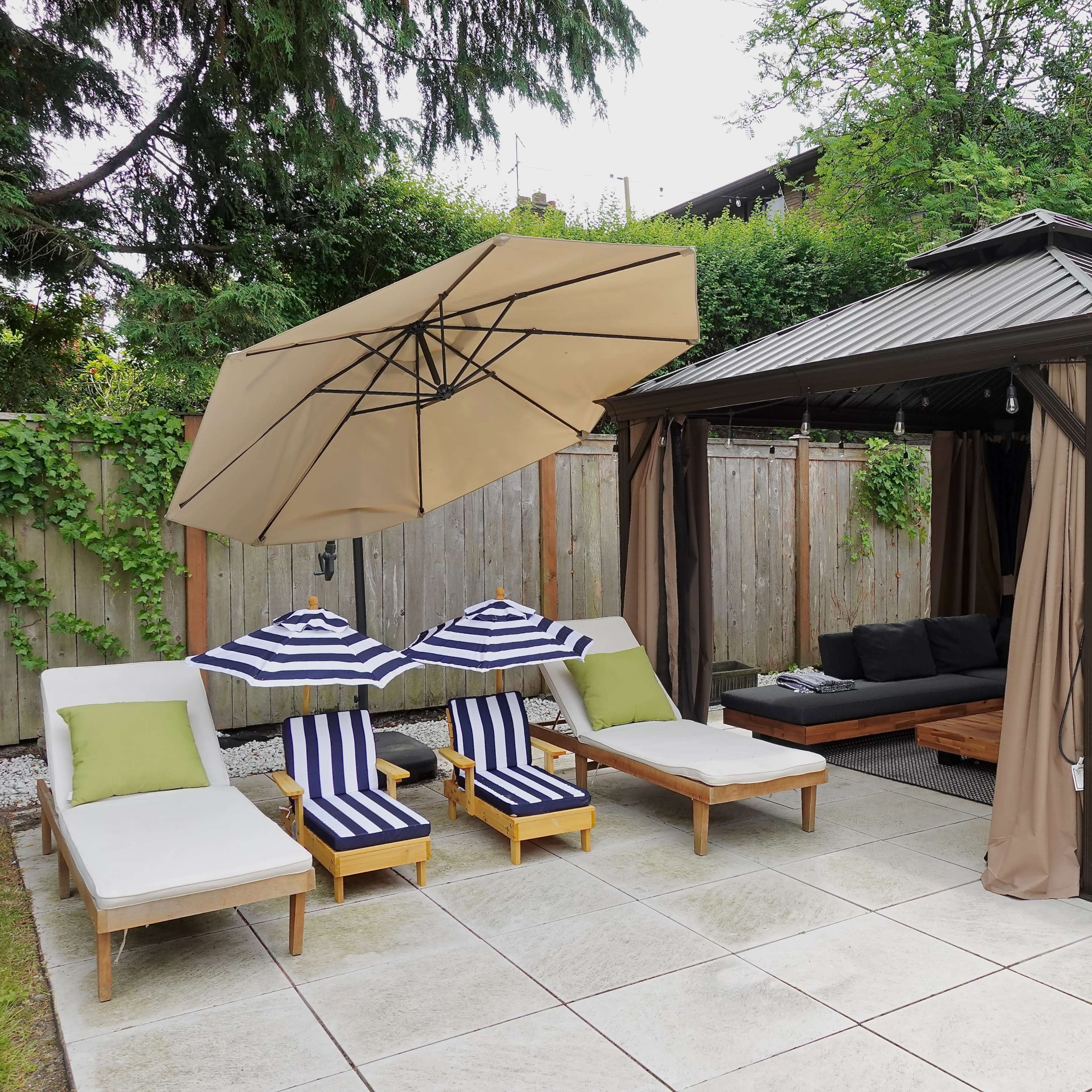 A patio area with two lounge chairs, a striped umbrella, and a gazebo adjacent to a grassy area surrounded by wooden fencing and greenery.